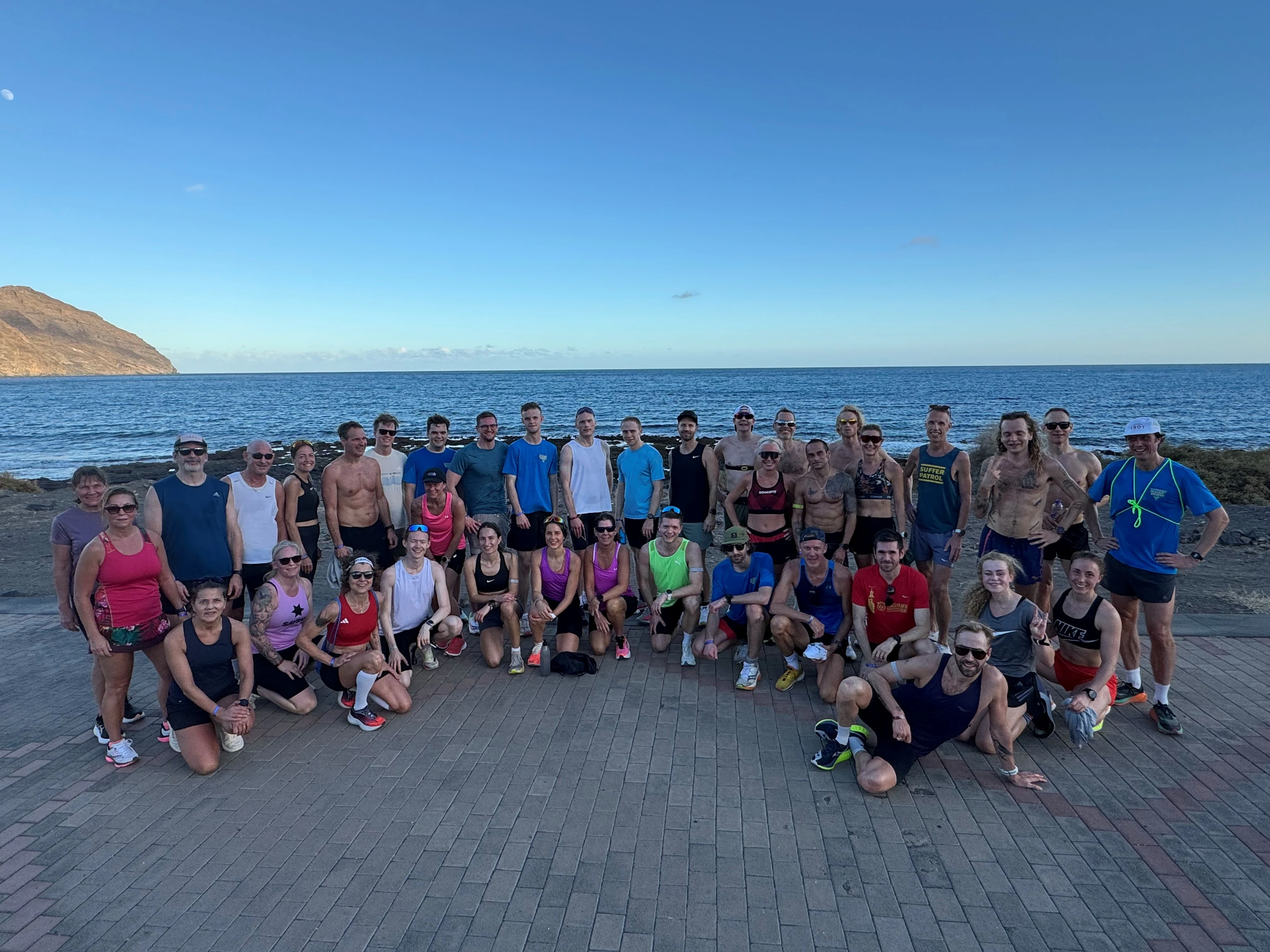 En stor grupp personer i träningskläder samlade vid havet på en stenlagd yta med hav och berg i bakgrunden