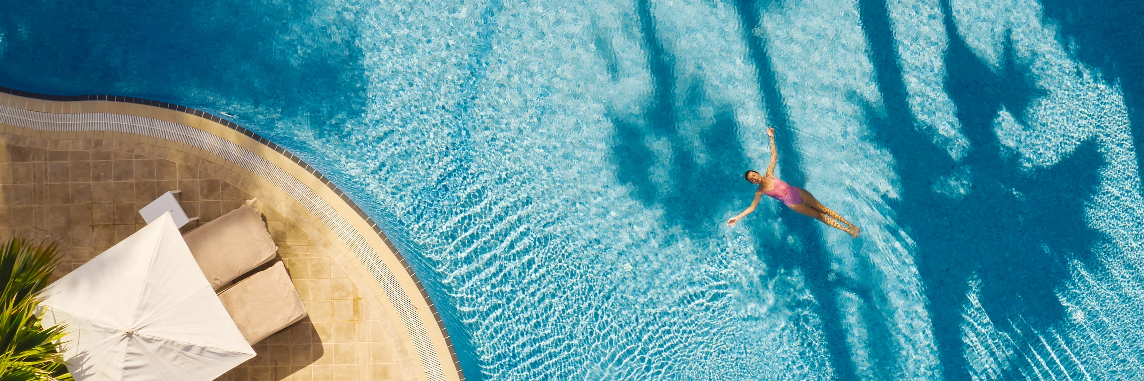 En person svømmer i en turkisblå pool set ovenfra, med solstole og parasoller ved poolkanten og palmeskygger, der falder hen over vandet.