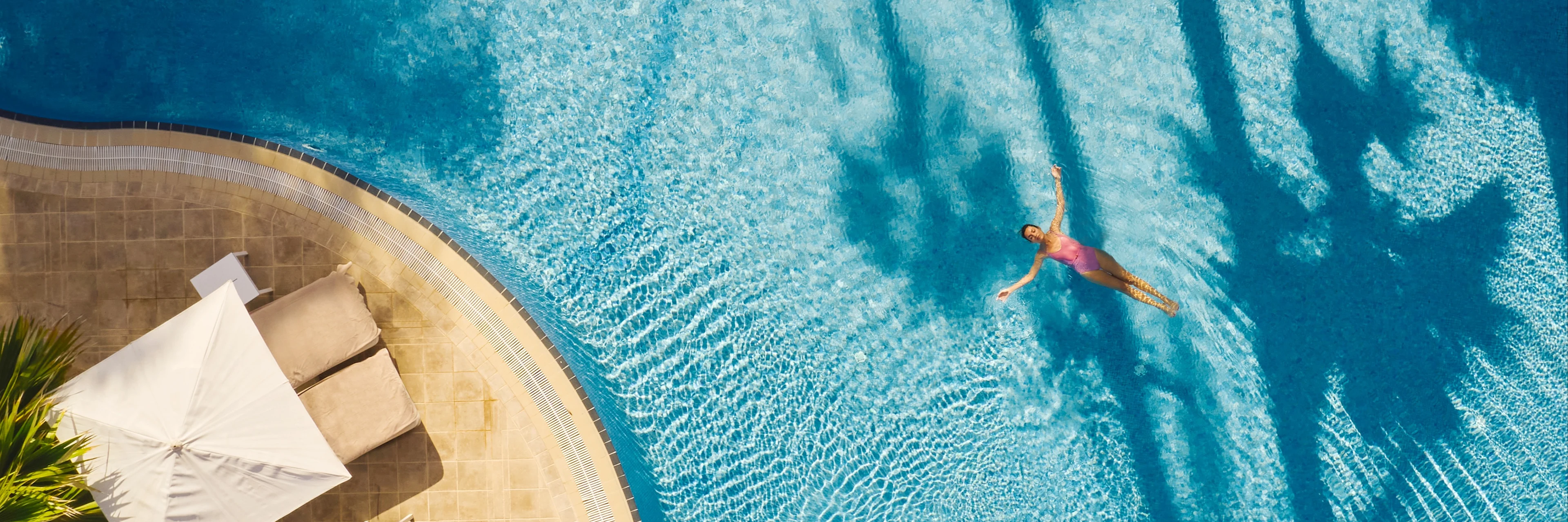 Henkilö ui turkoosinsinisessä uima-allas ylhäältä katsottuna, altaan reunalla on aurinkotuolit ja -varjoja ja palmujen varjot langeavat veteen.