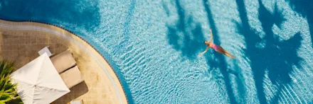 En person simmar i en turkosblå pool sedd uppifrån, med solstolar och parasoll vid poolkanten samt palmers skuggor som faller över vattnet.