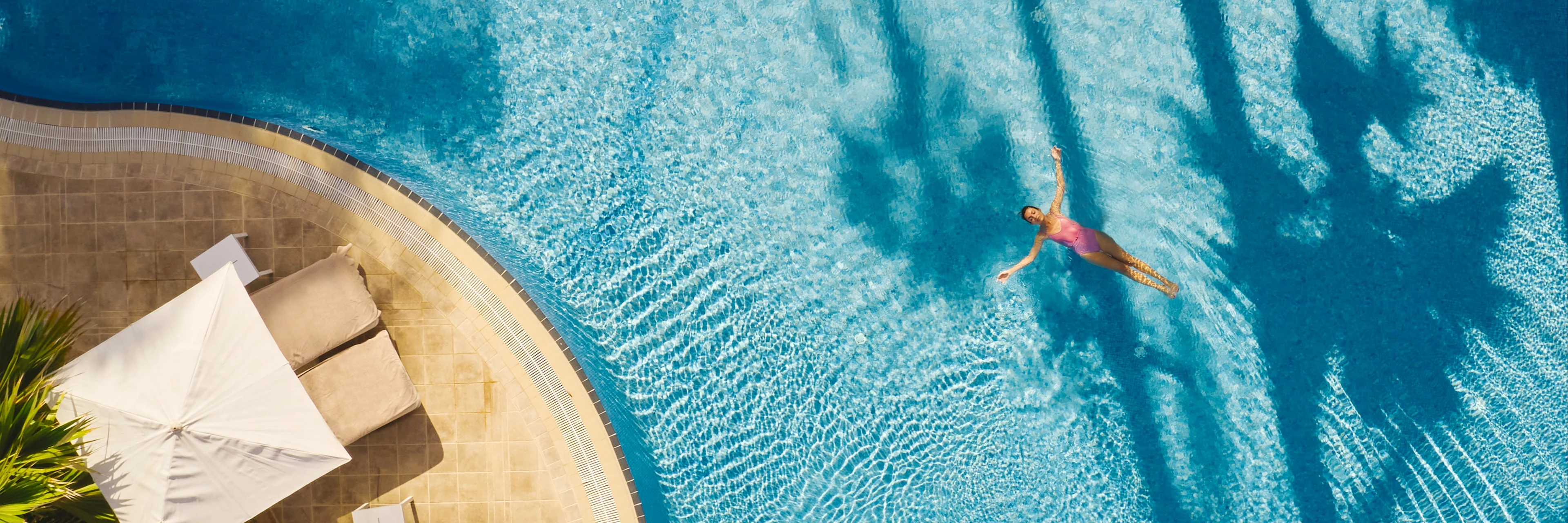 En person simmar i en turkosblå pool sedd uppifrån, med solstolar och parasoll vid poolkanten samt palmers skuggor som faller över vattnet.