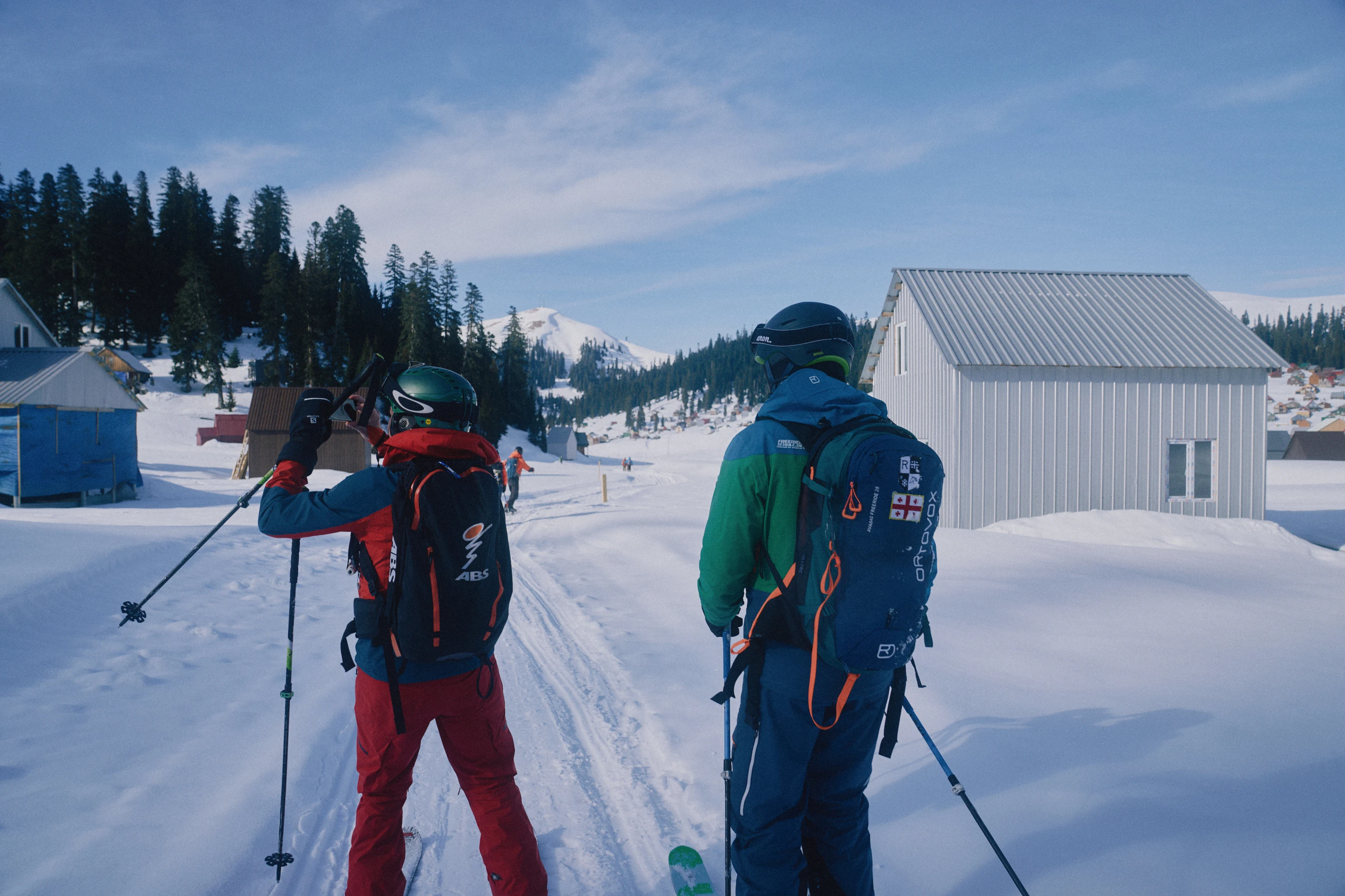 To skikjørere med ryggsekker går langs en snødekt vei i en vinterlandsby med fjell i bakgrunnen
