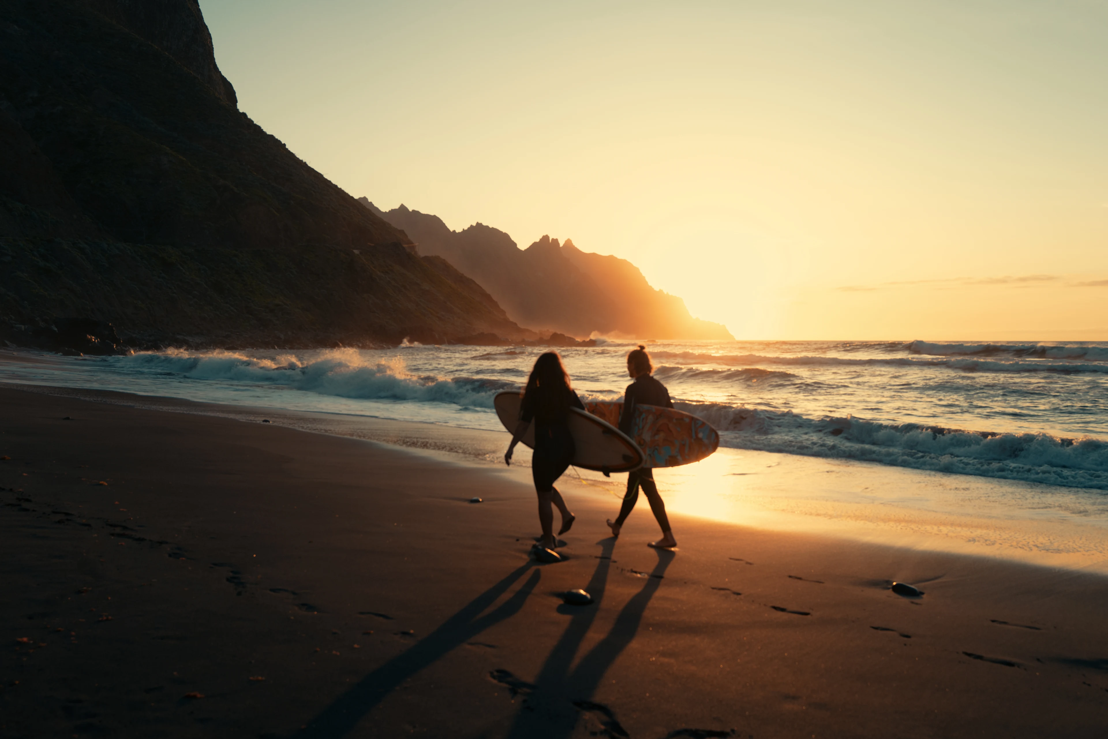 Två surfare promenerar längs en svart sandstrand på Teneriffa med Atlantens vågor och bergiga kustlandskap i bakgrunden.