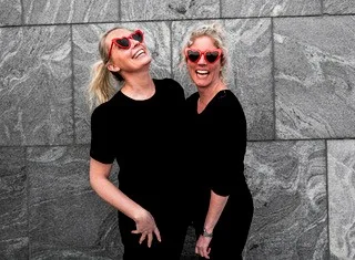Profilbilde av Kine og Elisabeth som står sammen og ler ikledd sort med røde hjerteformede solbriller på. De står foran en vegg med grå fliser. 