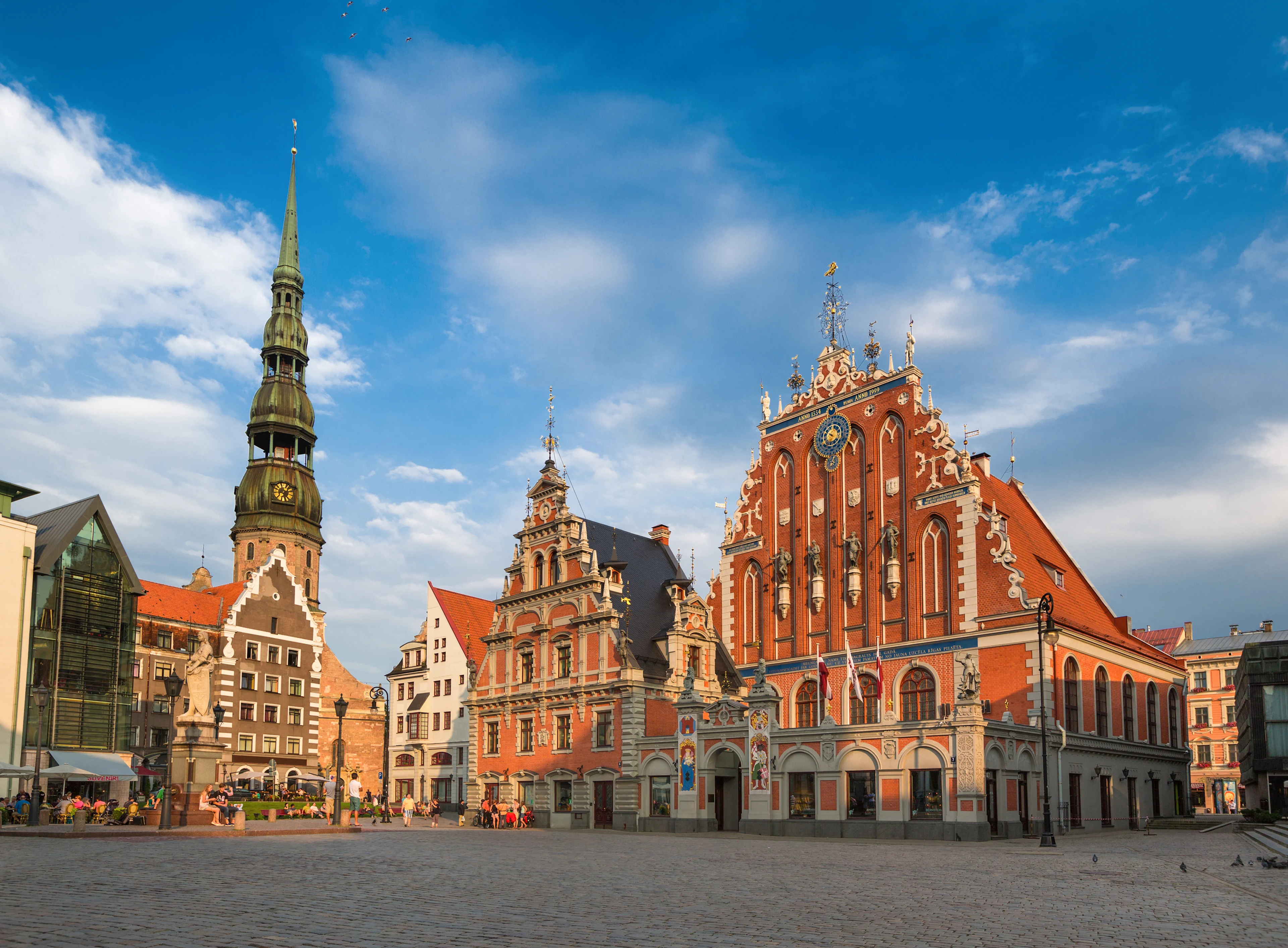Et foto fra pladsen i den gamle bydel i Riga med gamle smukke bygninger og en stor kirke med et højt tårn. Alt sammen under en blå himmel.
