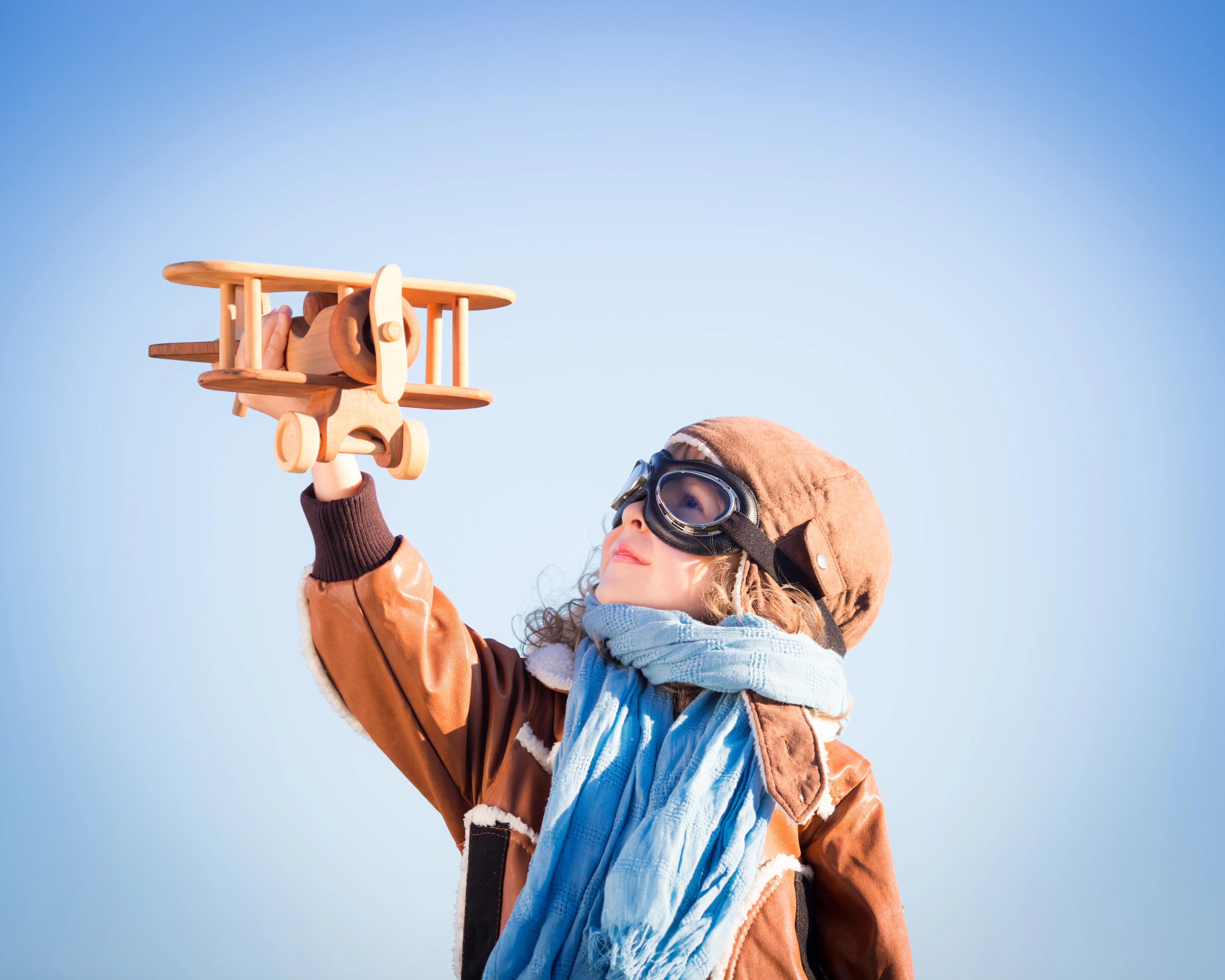En ung flicka klädd som en pilot med en brun läderjacka, flygarhuva och skyddsglasögon håller upp ett träflygplan mot en klarblå himmel. Hon bär också en ljusblå halsduk och ser inspirerad ut.