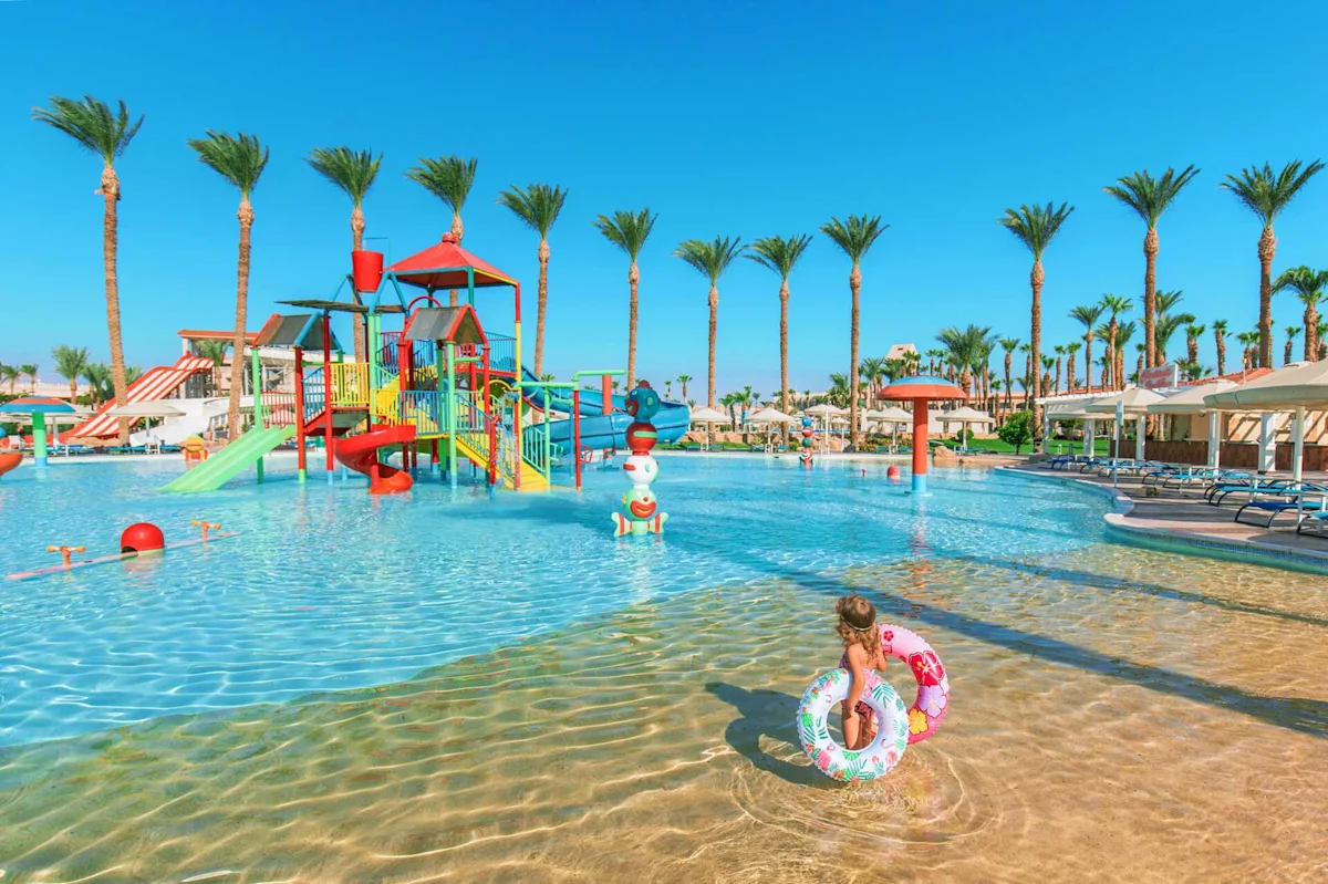 Ett färgstarkt vattenland med rutschkanor och palmer runtomkring, där ett barn leker i det grunda, klara vattnet.