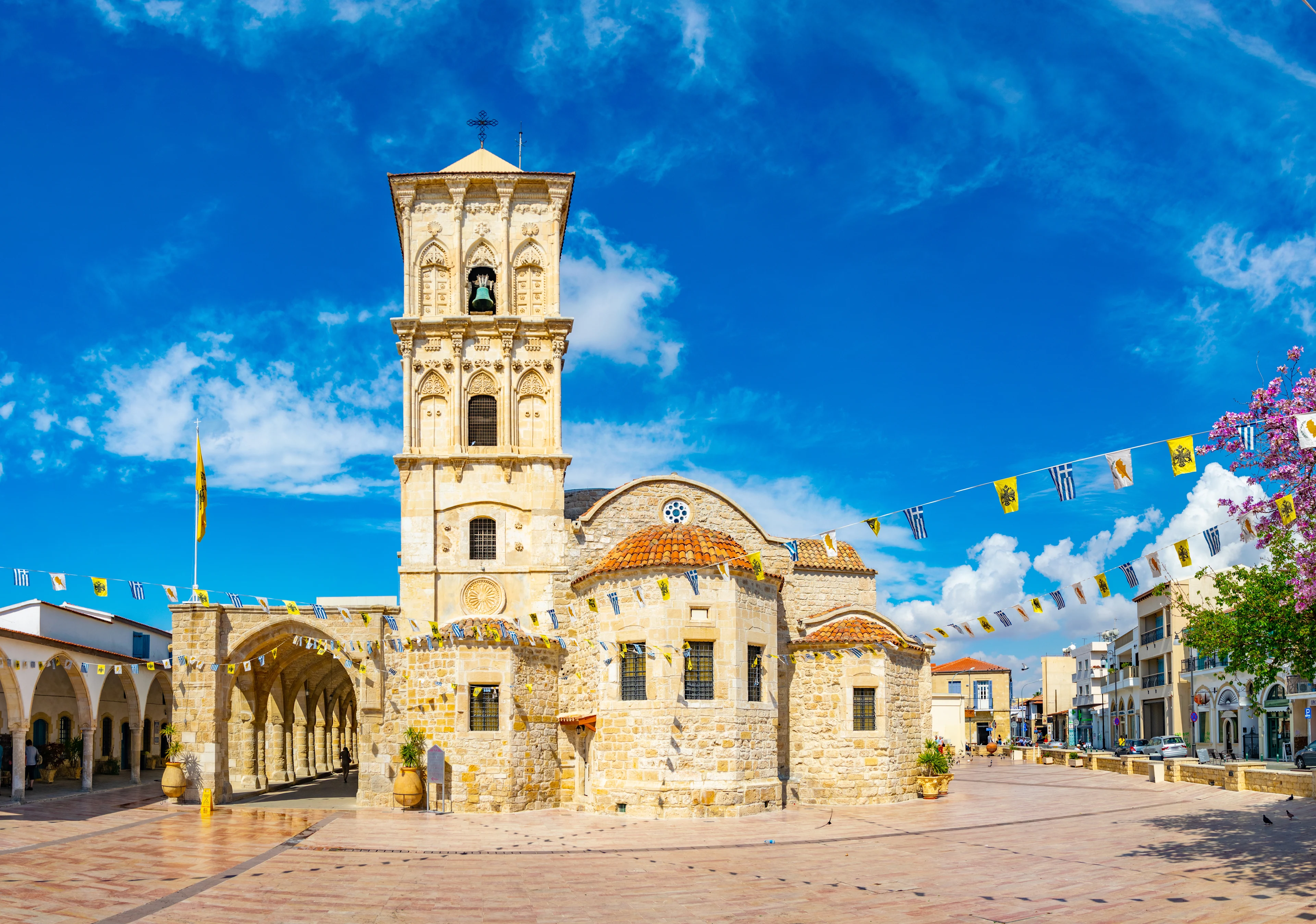 Sankt Lazarus-kyrkan i Larnaca med traditionell stenarkitektur och färgglada flaggor under en blå himmel.