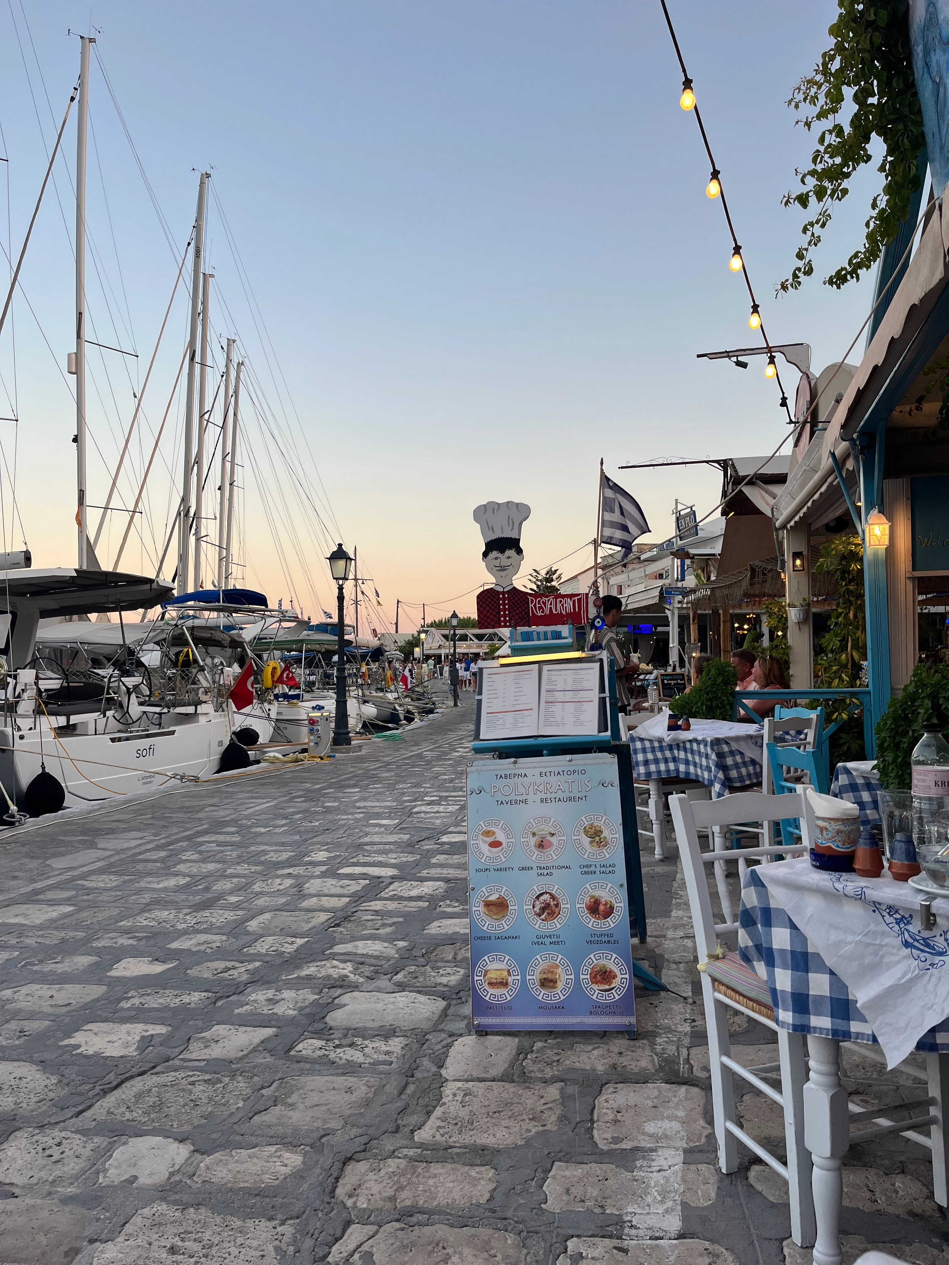 Solnedgång vid hamnpromenad med segelbåtar, uteservering, meny och en kockstaty – en inbjudande kvällsstämning på Samos.