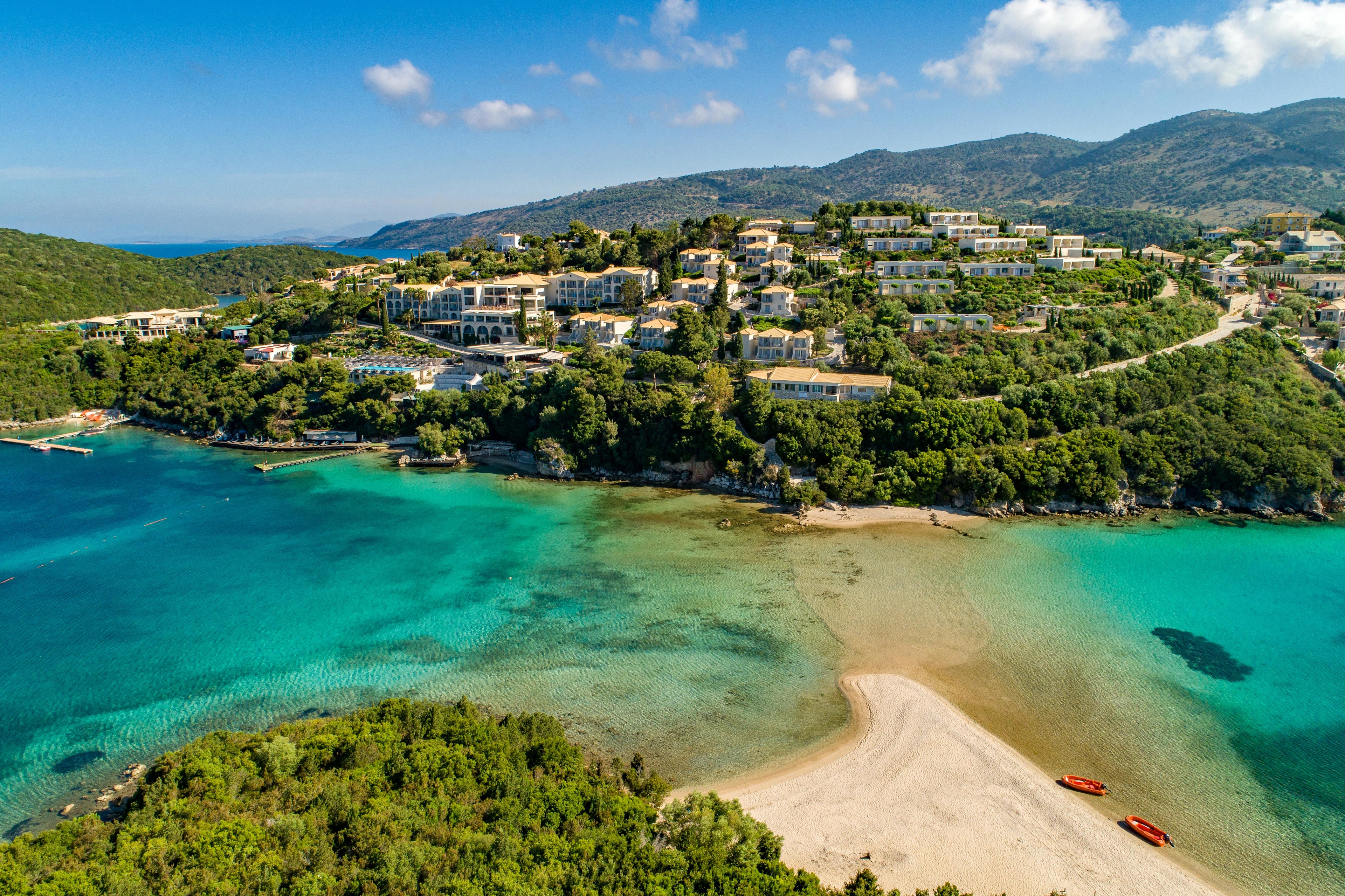 Luftfoto af en kyst med turkisblåt vand, strand og et resort omgivet af grønne områder.