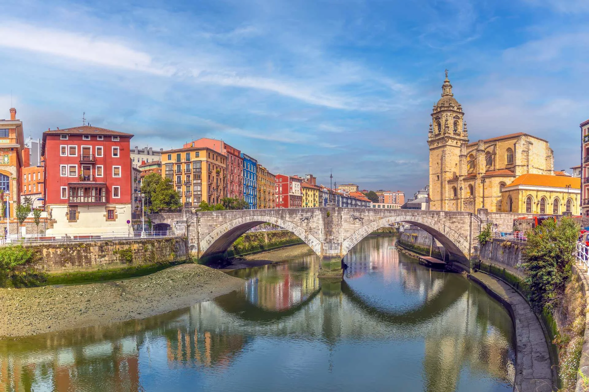 Färgglada byggnader och den historiska San Antón-bron över floden Nervión i Bilbao, med San Antón-kyrkan i bakgrunden under en klarblå himmel."