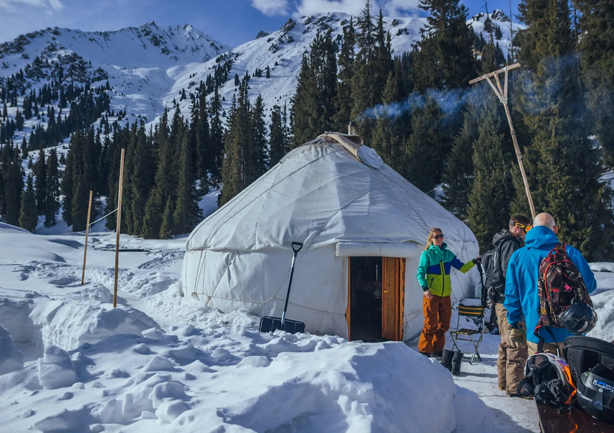 Ett runt tält står i snön omgivet av granar och berg, medan flera personer med packning samlas utanför.