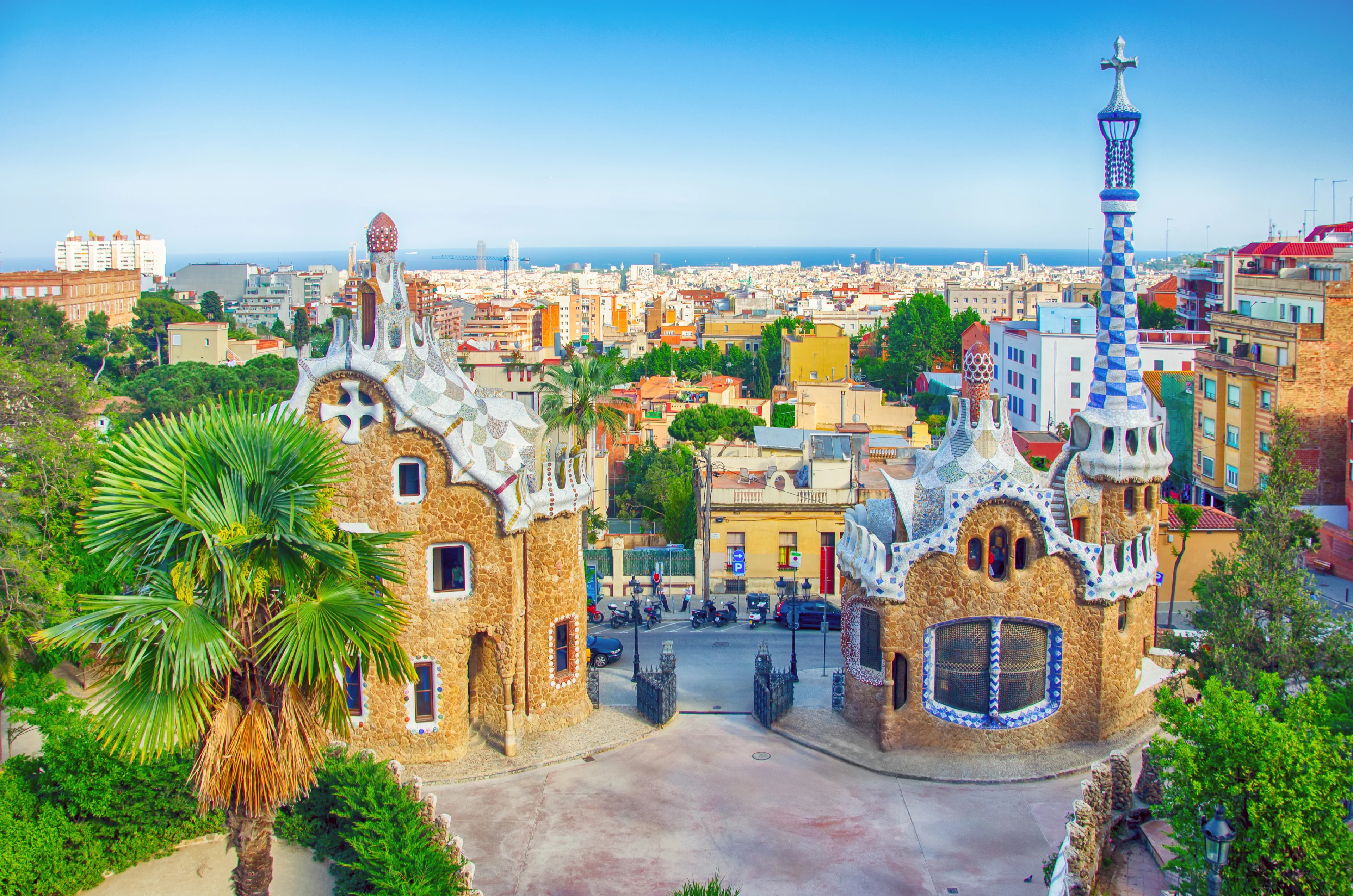 Färgglada byggnader med mosaikfasader i Gaudís karakteristiska arkitektoniska stil i Park Güell, Barcelona. Stadens silhuett och Medelhavet skymtas i bakgrunden, omgivna av frodig grönska och palmer.