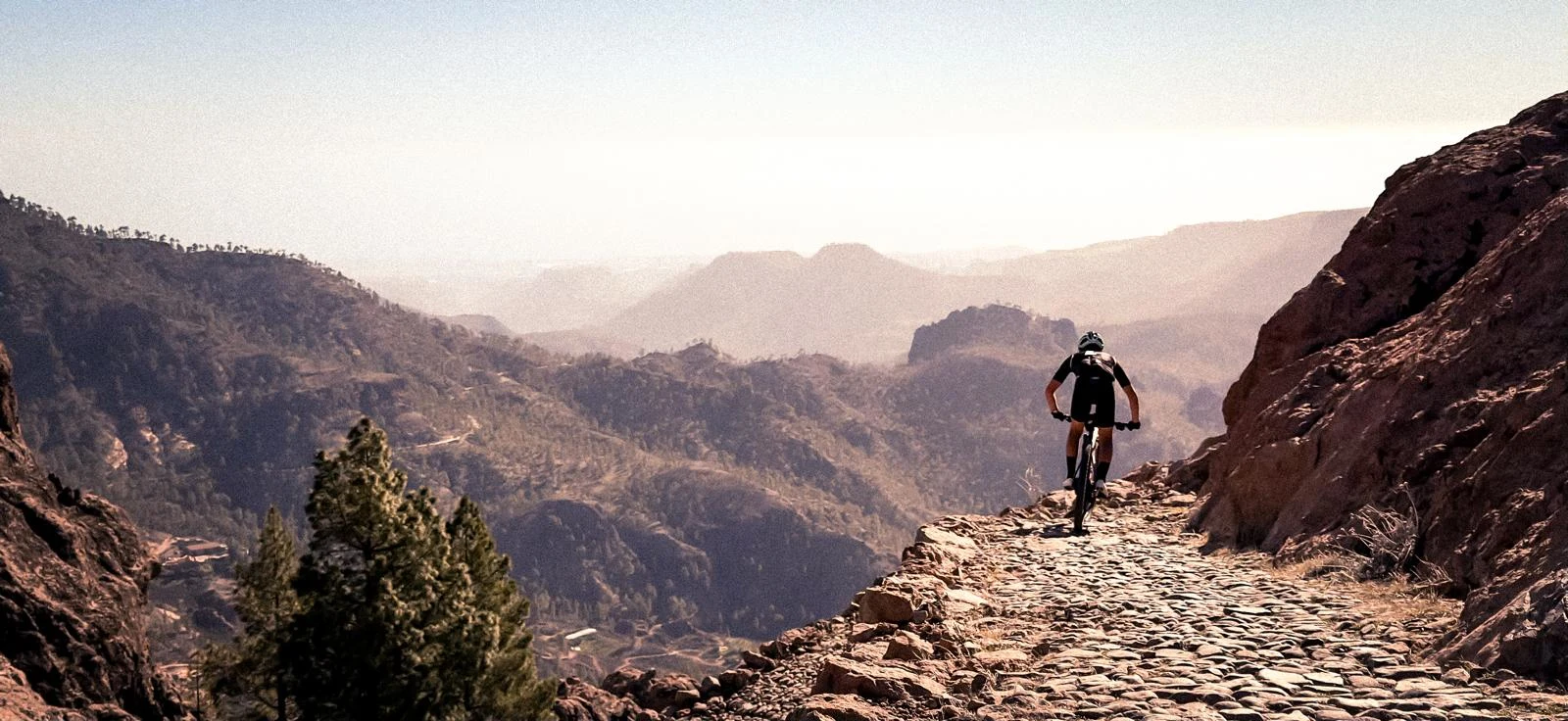 En cyklist färdas på en smal stenstig högt uppe i ett bergsområde, med en vidsträckt vy över dalar och berg i bakgrunden