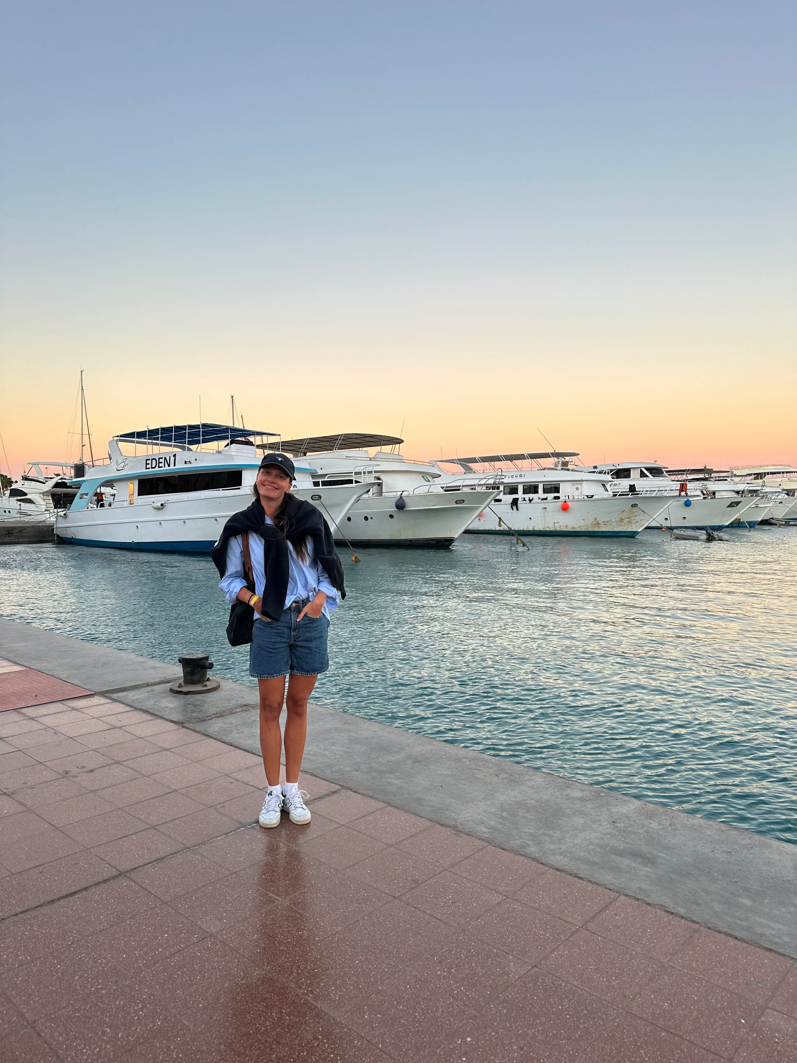 Anja som står på en brygge med yachetr i bakgrunnen. Hun er kledd i shorts og skjorte med mørk blå genser over skuldrene og caps 