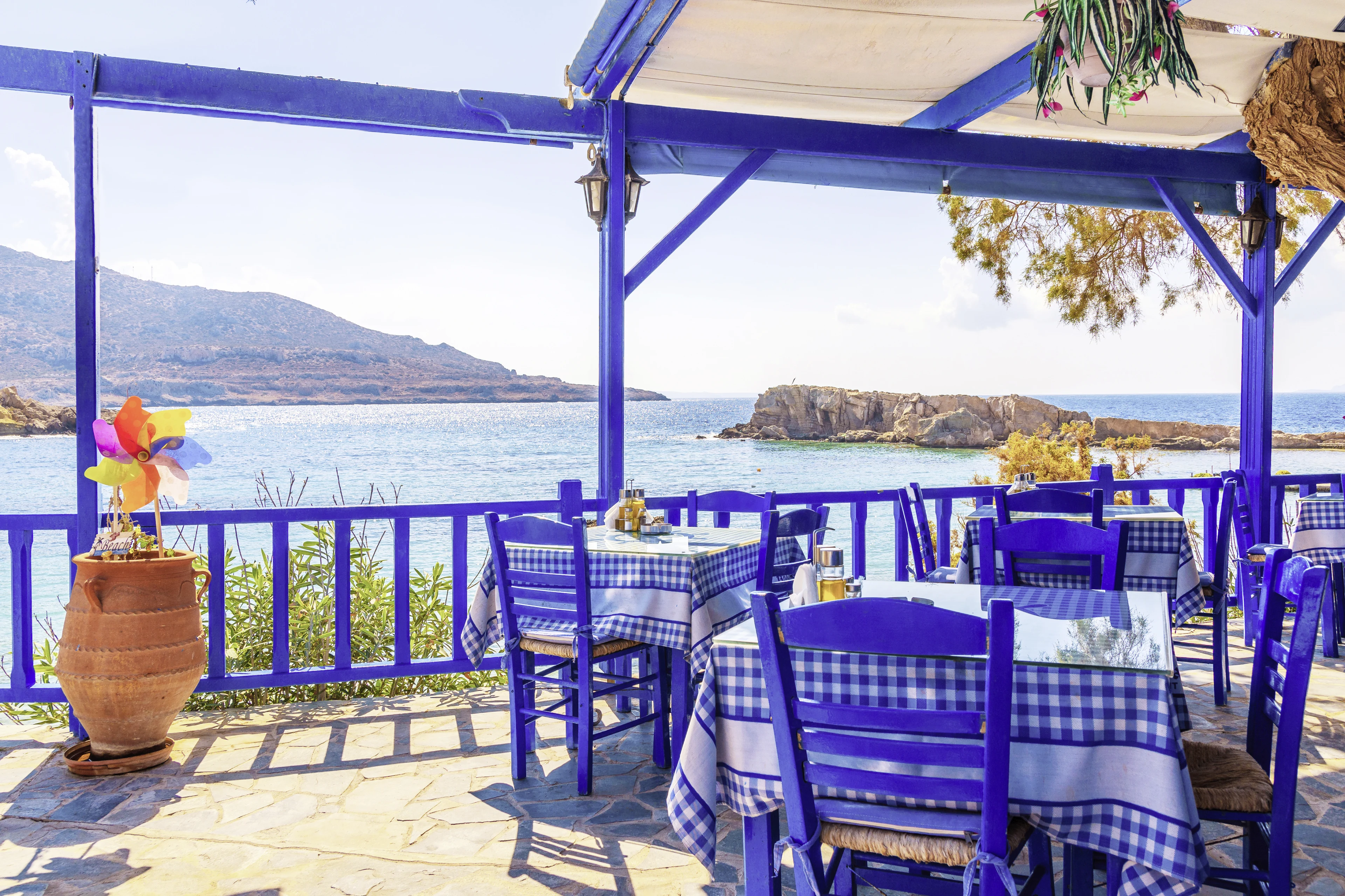 Restaurant med blåa stolar och bord med klassiskt blå-vitrutiga dukar. Havet och bergen i bakgrunden.