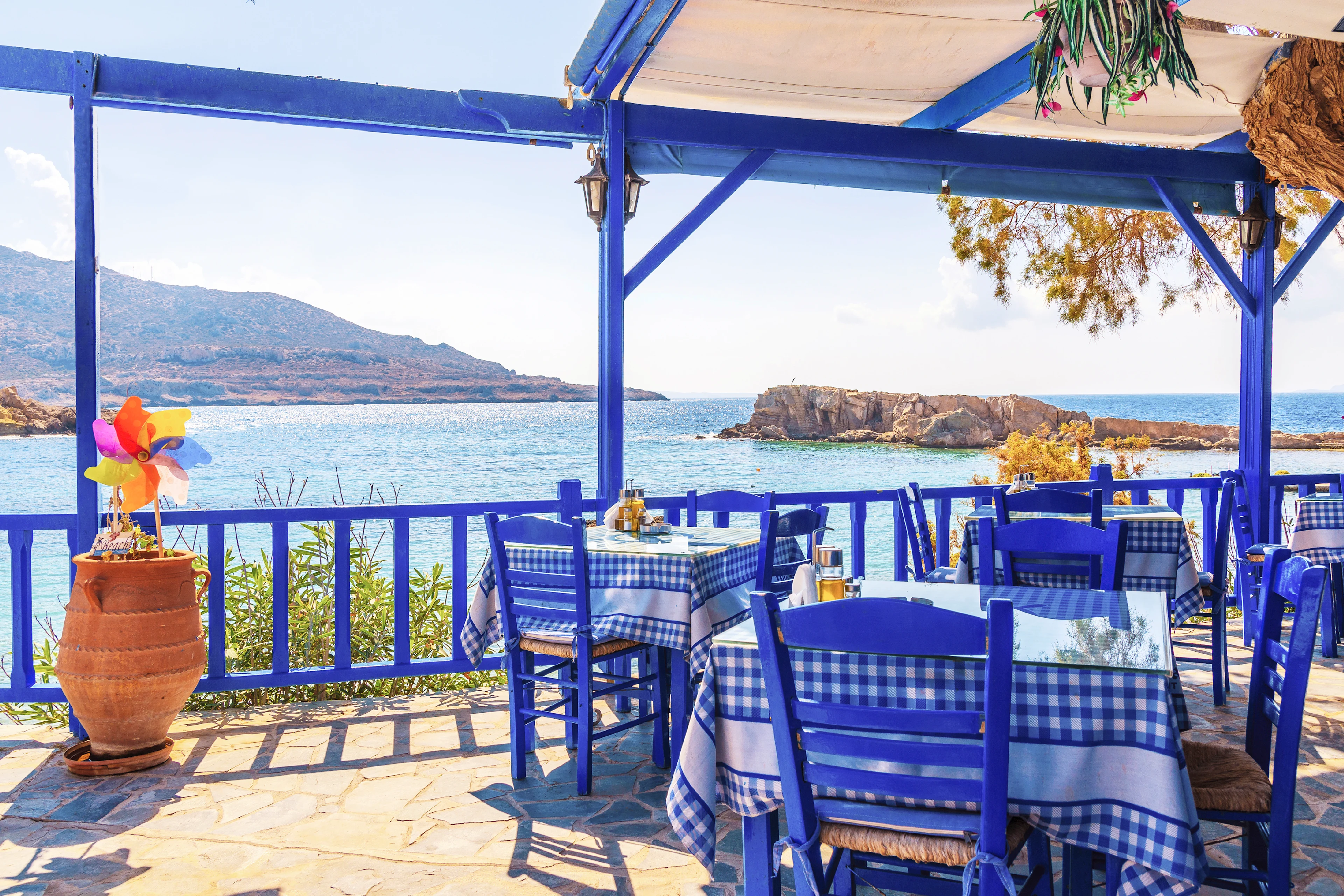 Restaurant med blåa stolar och bord med klassiskt blå-vitrutiga dukar. Havet och bergen i bakgrunden.