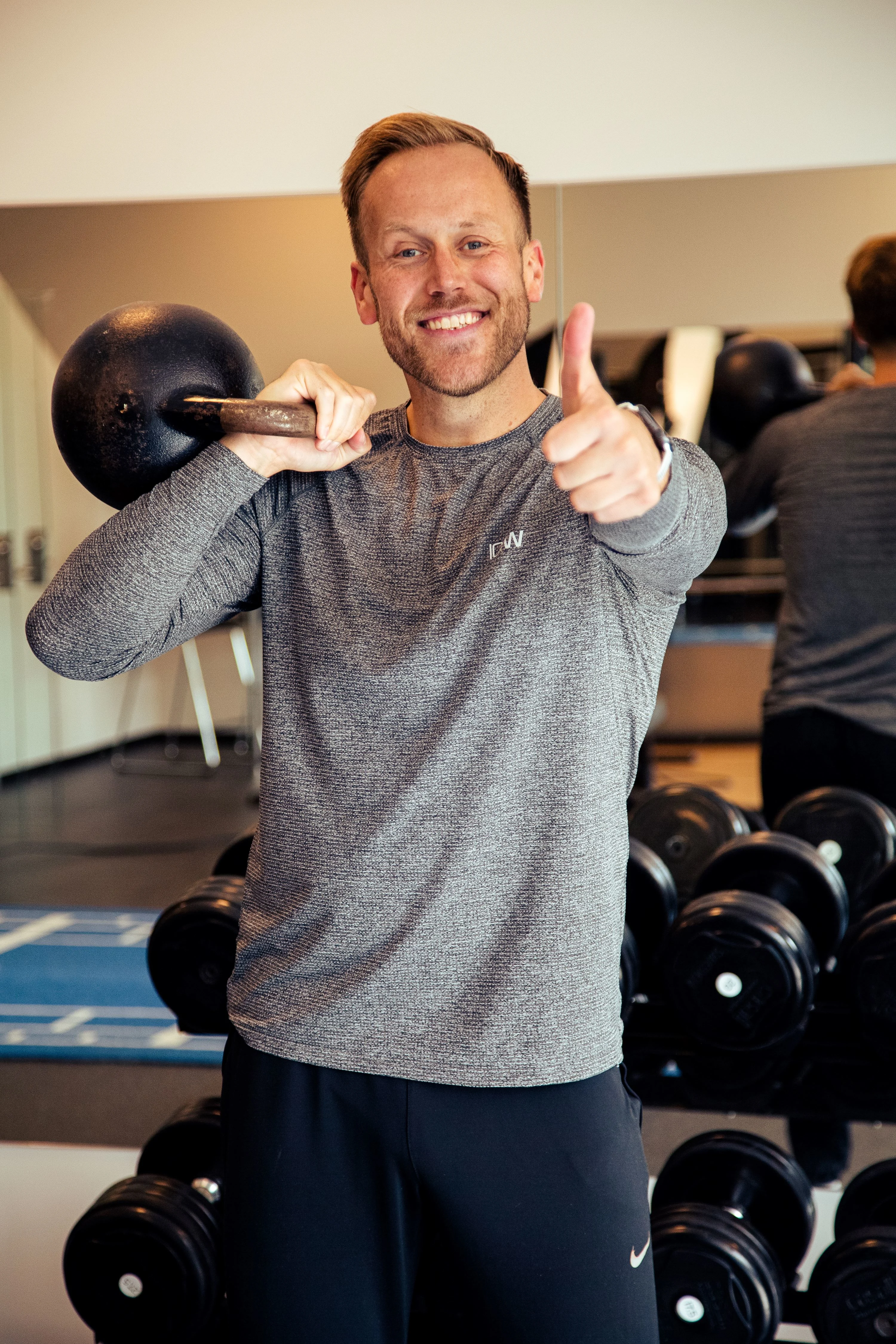 En man i en grå långärmad tröja med en svart kettlebell i ena handen över axeln.