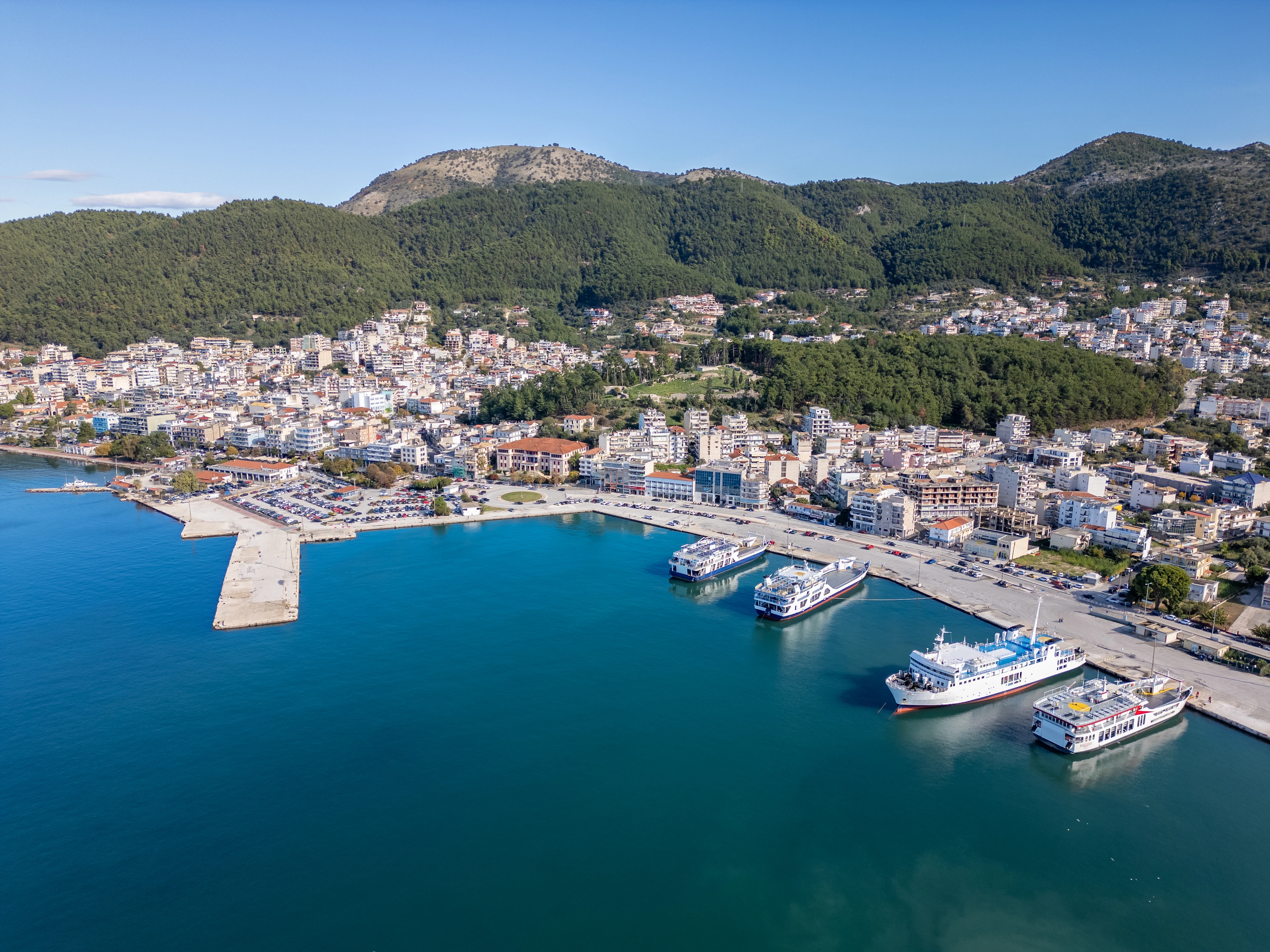 Flygfoto över hamnen i Igoumenitsa med färjor vid kajen och grönskande berg i bakgrunden.