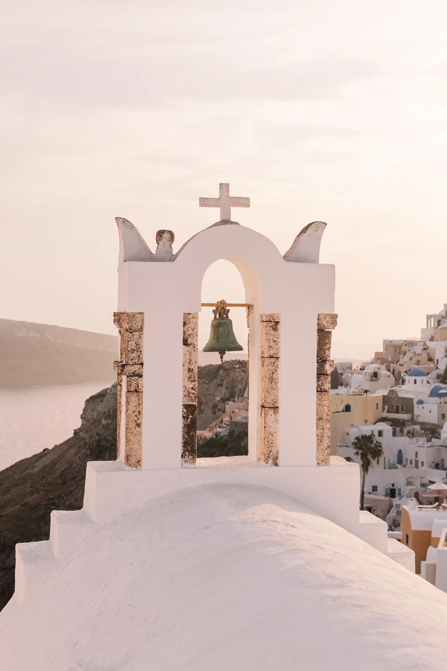 Hvit kirkeklokke med kors på en ås i Santorini, med utsikt over de ikoniske hvite og blå bygningene og Egeerhavet i solnedgangen.