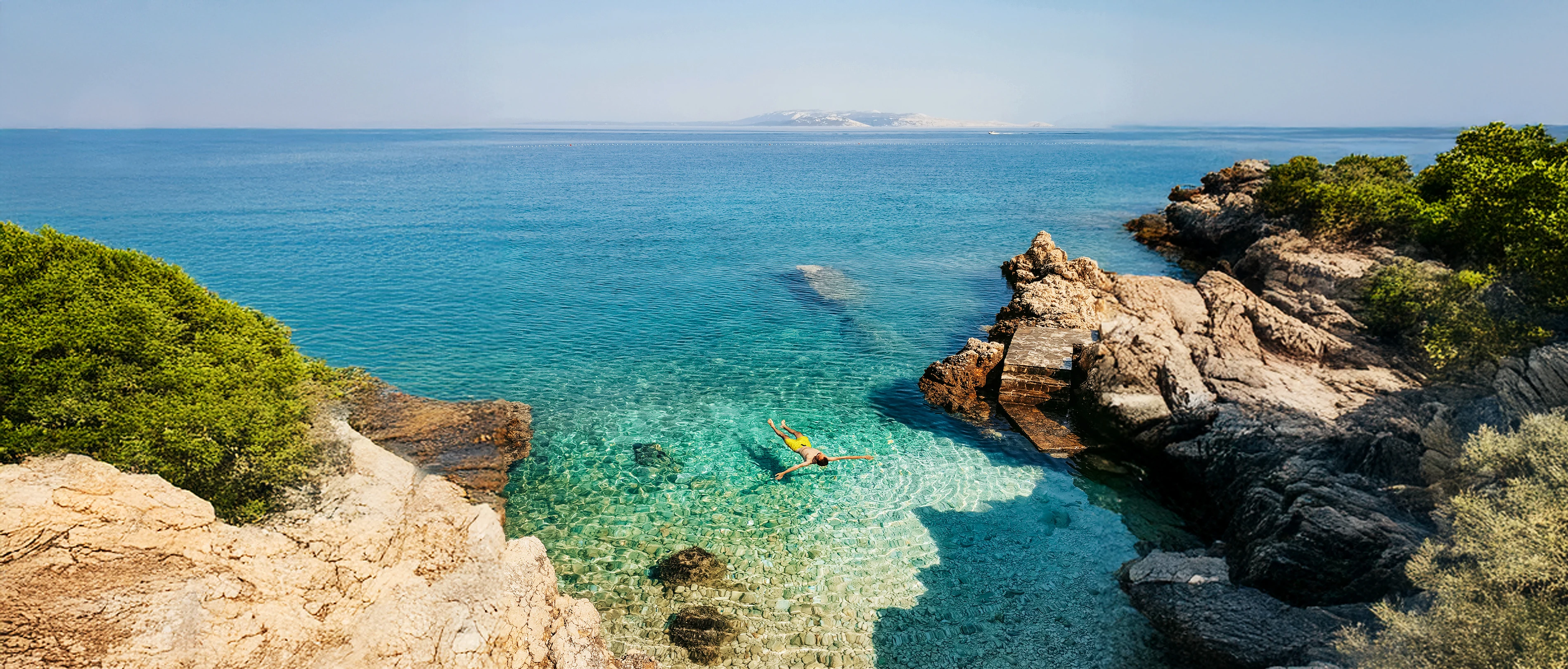Person som flyter i klart turkost vatten vid en liten stenig vik omgiven av klippor och grönska under en solig dag.