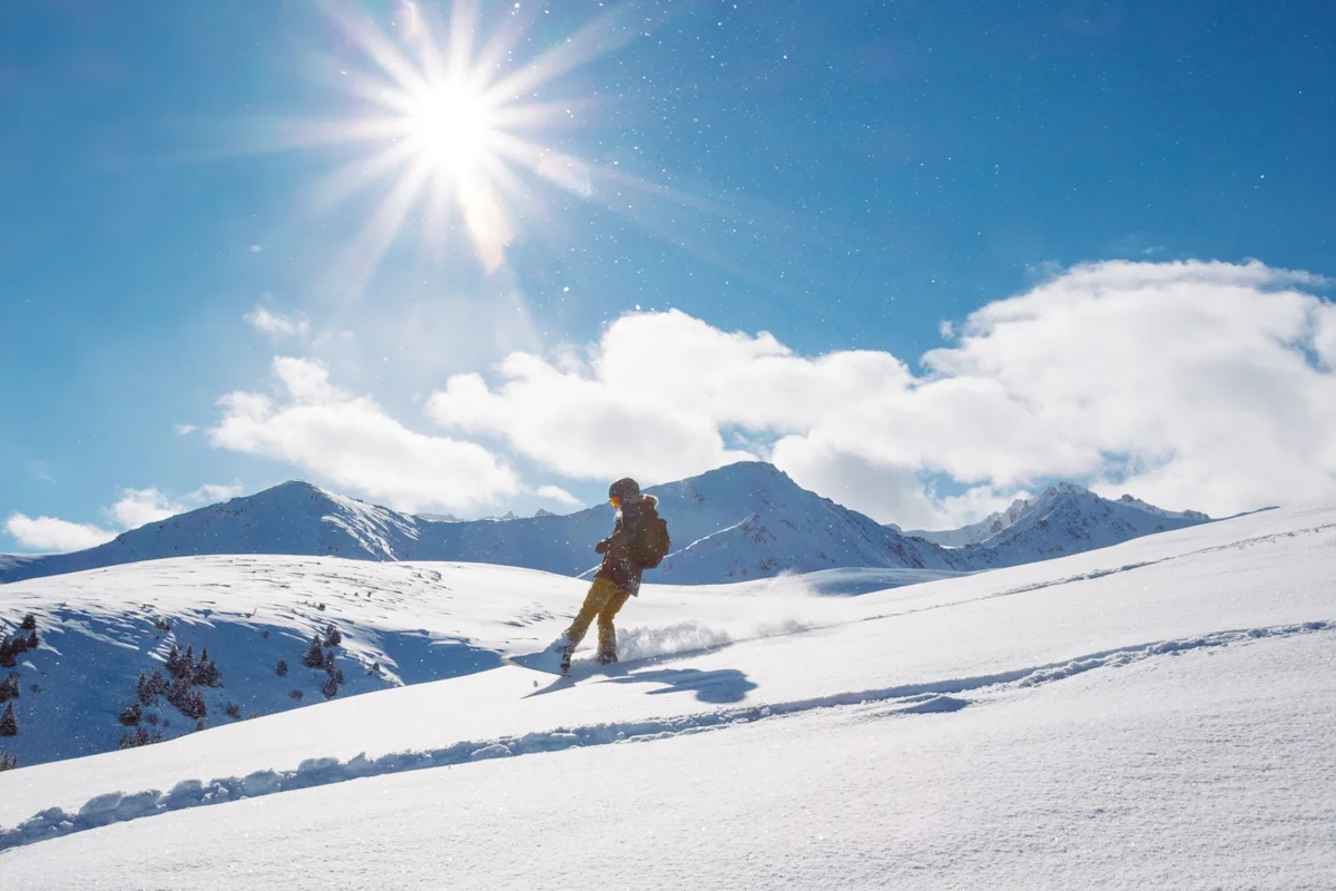 En person rör sig genom ett snötäckt bergslandskap i starkt solsken, med höga toppar och blå himmel i bakgrunden.