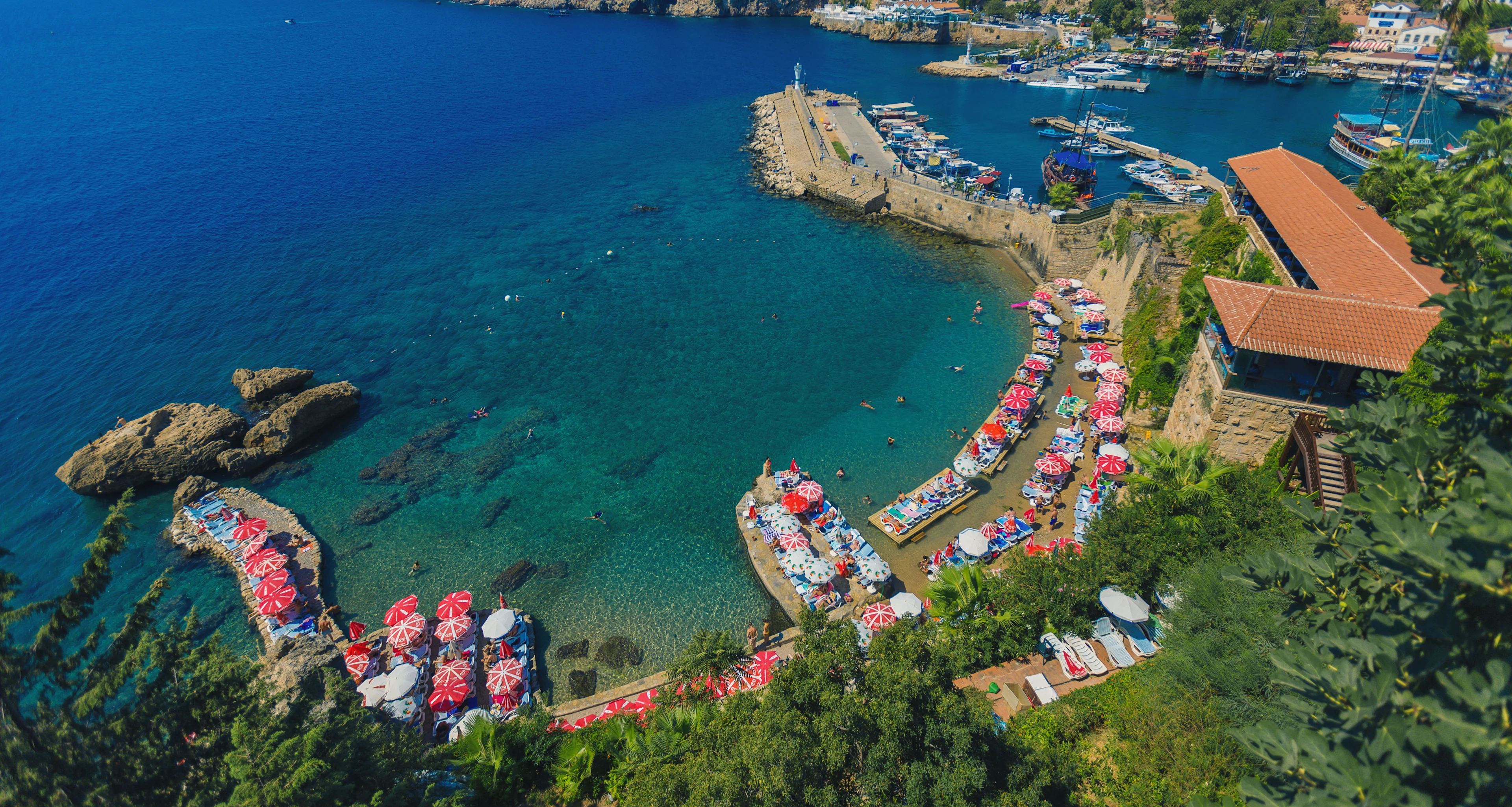 Flygfoto över en skyddad vik med klart turkost vatten, solstolar och parasoller och en småbåtshamn i Antalya