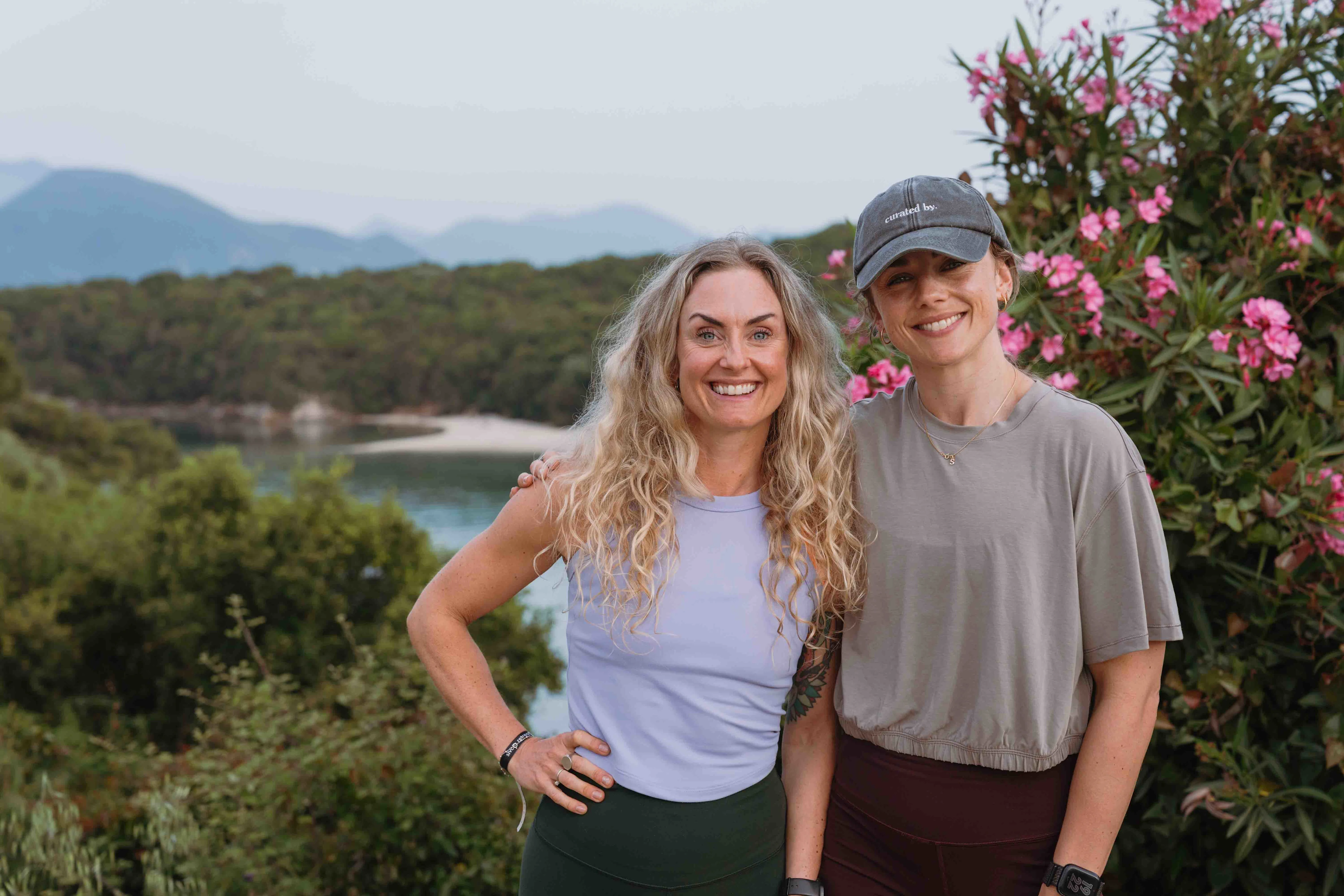 Silje Thorstensen og Pia Seeberg står sammen ute foran en oleander i blomst med grønn skog og hav i bakgrunnen.
