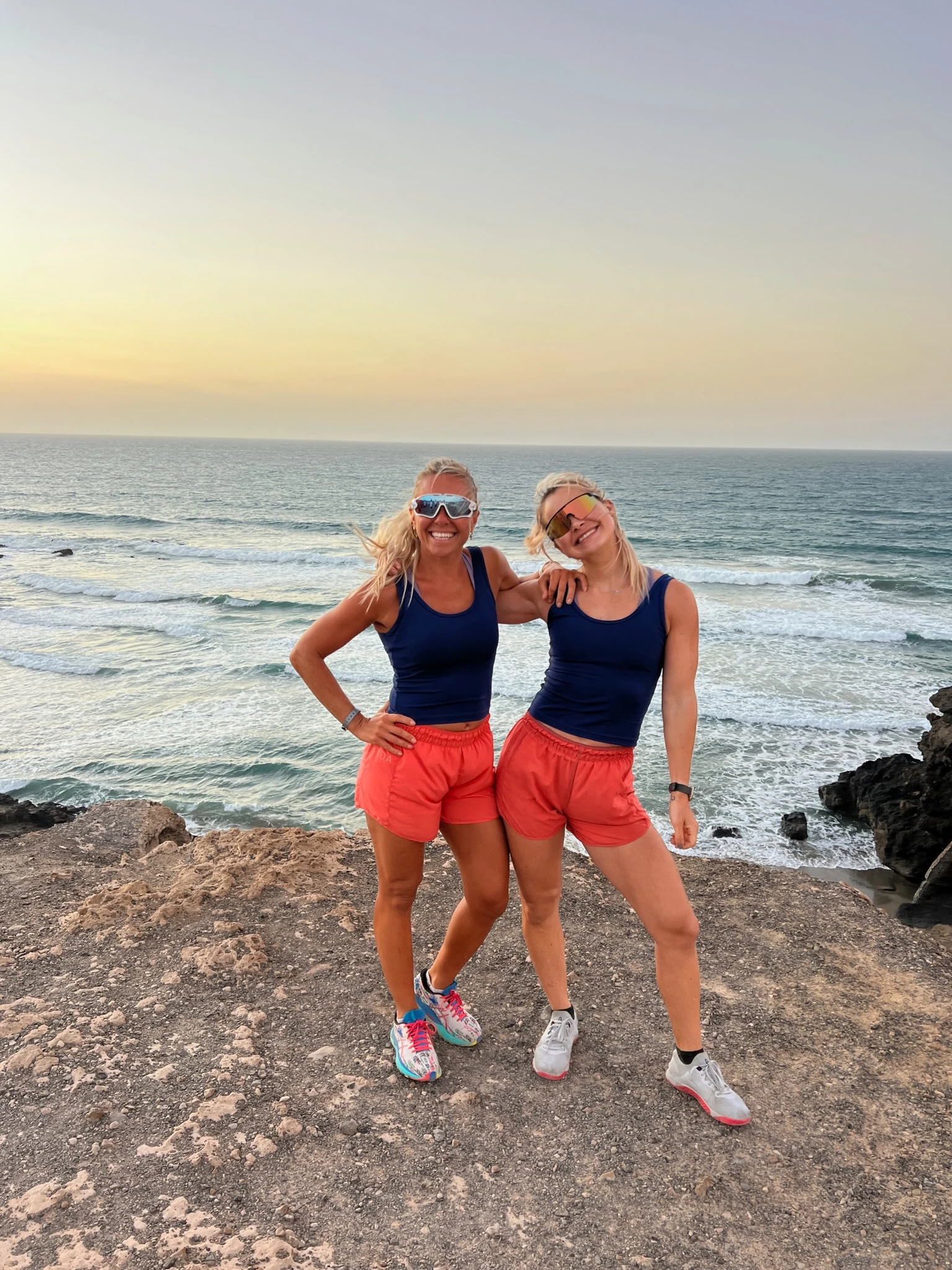 Charlotte og Camille står på en strand og holder hverandre på skulderen likt kledd i blå singel og rød schorts.