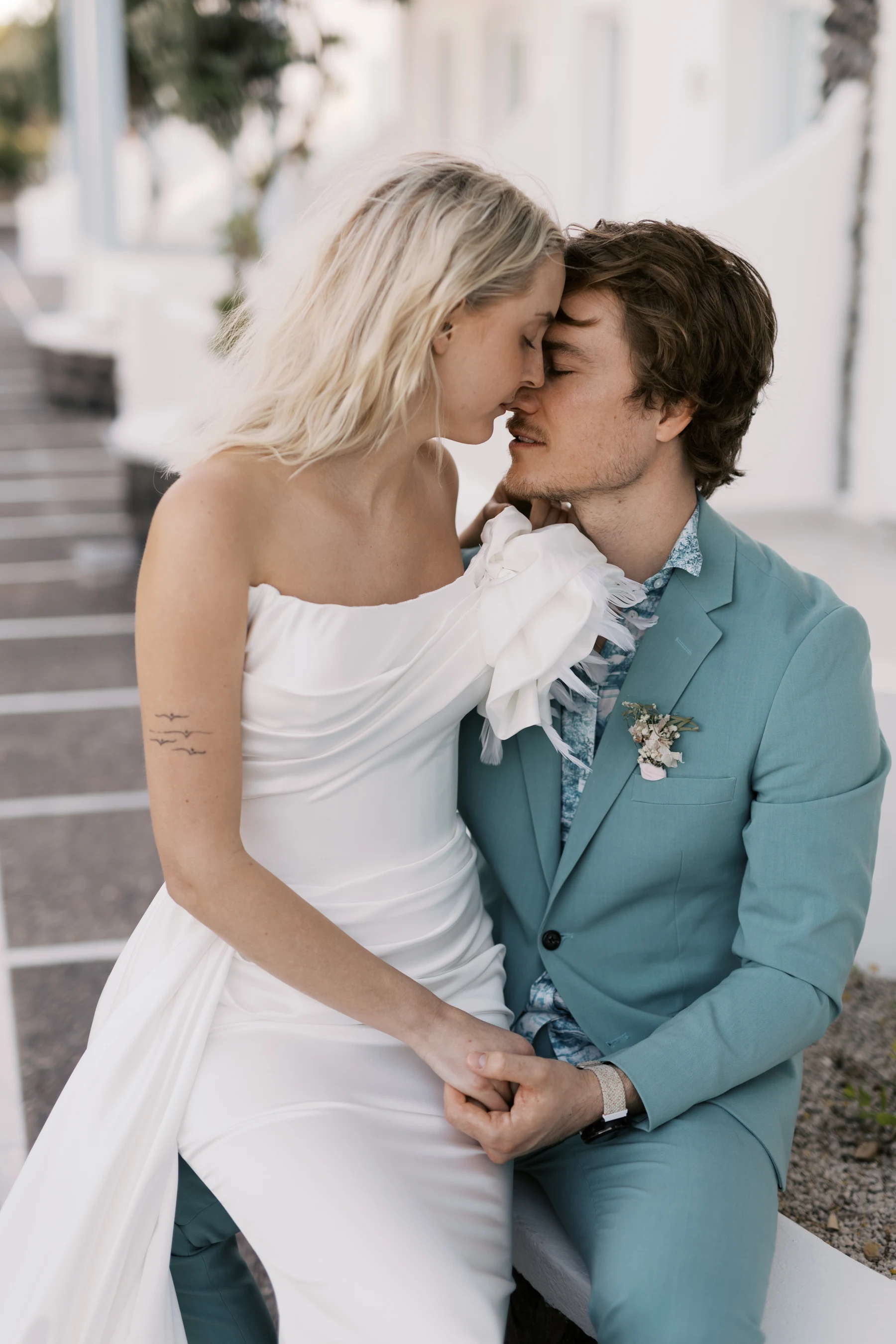 Nygift par deler et intimt øyeblikk på Santorini. Bruden i en elegant hvit kjole og brudgommen i en turkis dress sitter tett sammen, holder hender og lener seg inn for en kjærlig berøring.