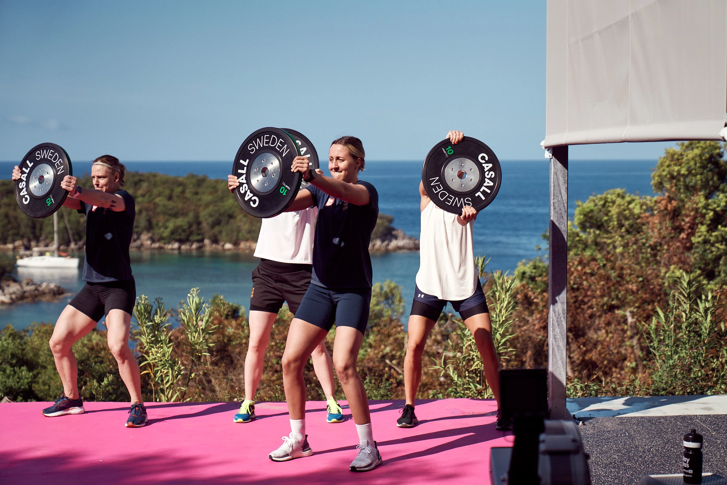 Ryhmä harjoittelee painolevyjen kanssa ulkotreenausalustalla, jolta on näkymä merelle ja vihreille rinteille Sivotassa, Kreikassa.
