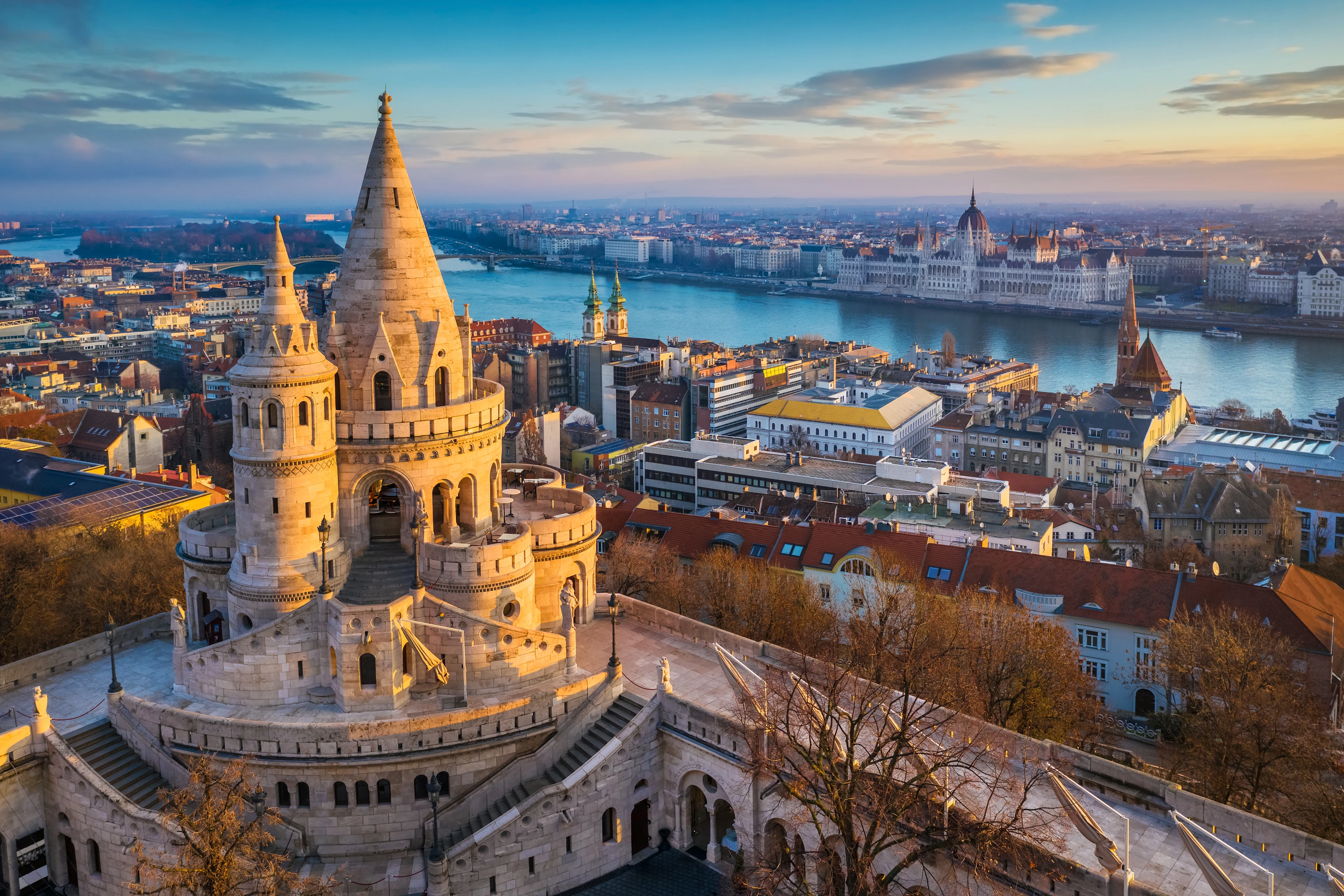 Panoramautsikt över Budapest med Fiskarbastionens torn i förgrunden och floden Donau som flyter genom staden. På andra sidan vattnet syns Ungerns parlament, omgivet av historiska byggnader i det varma kvällsljuset.