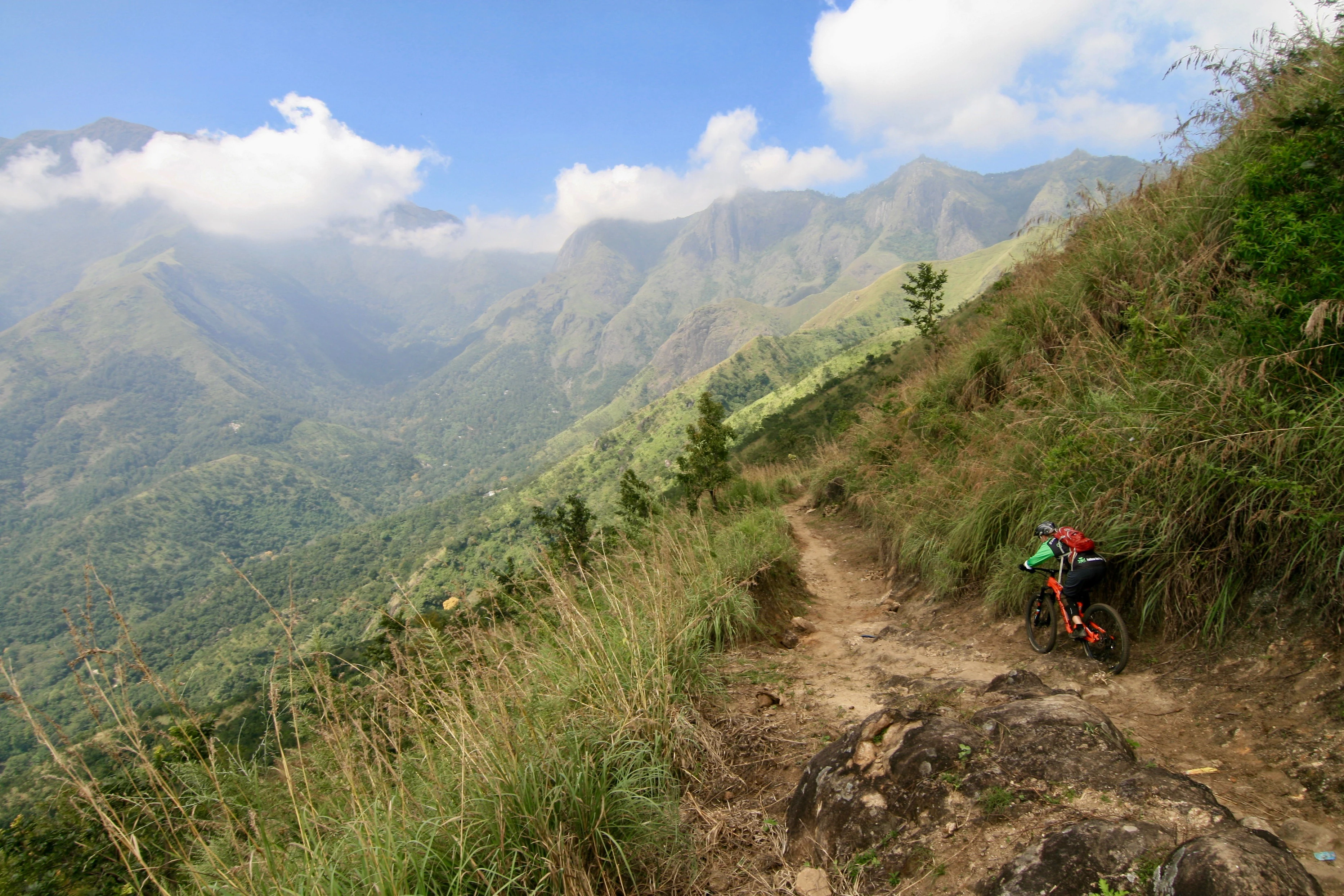 Cyklist som färdas på en smal bergsstig omgiven av grönskande sluttningar och dimhöljda berg