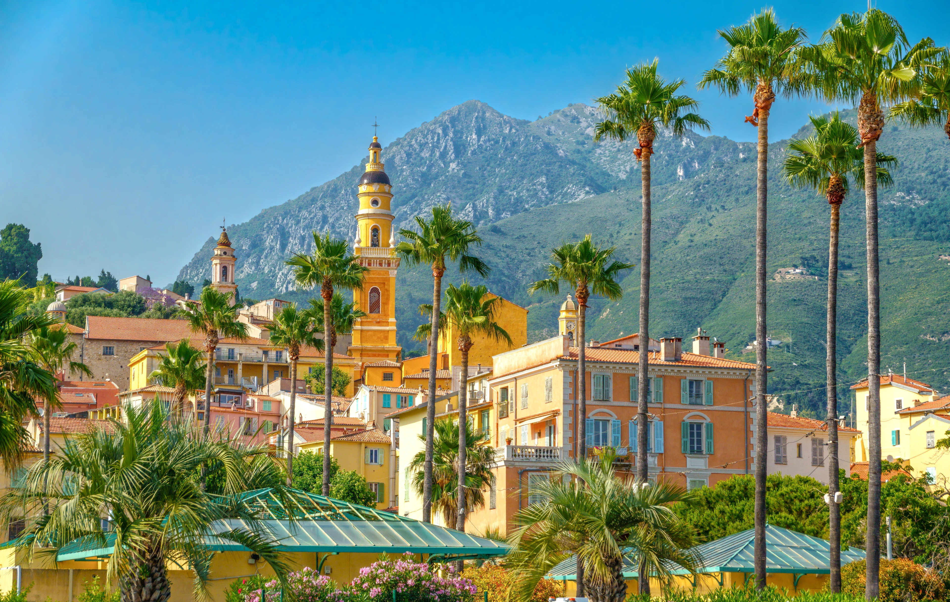 Menton på Franska rivieran med färgglada hus, palmer och kyrktorn mot de gröna bergen i bakgrunden.