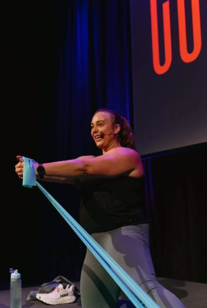 Profilbilde: Lotte Skullerud som instruerer kledd i sort topp og grå bukser og trekker i en lys blå strikk.