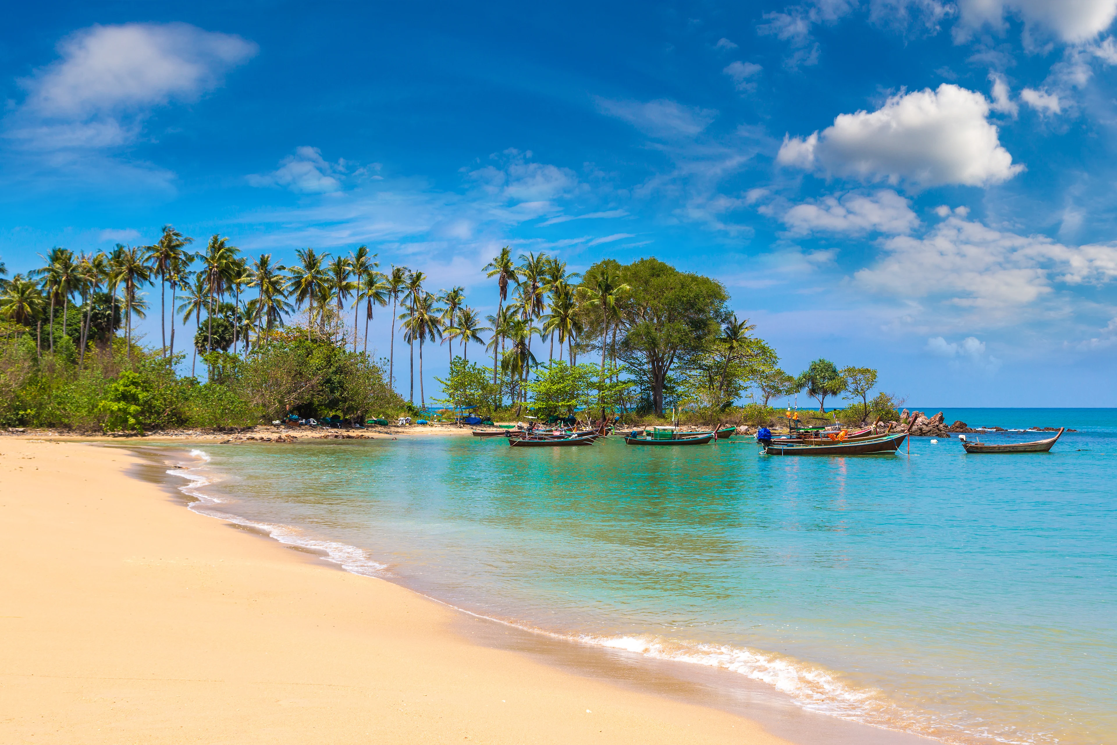 Sandstrand med turkost vatten, traditionella longtailbåtar och vajande palmer på Koh Lanta.
