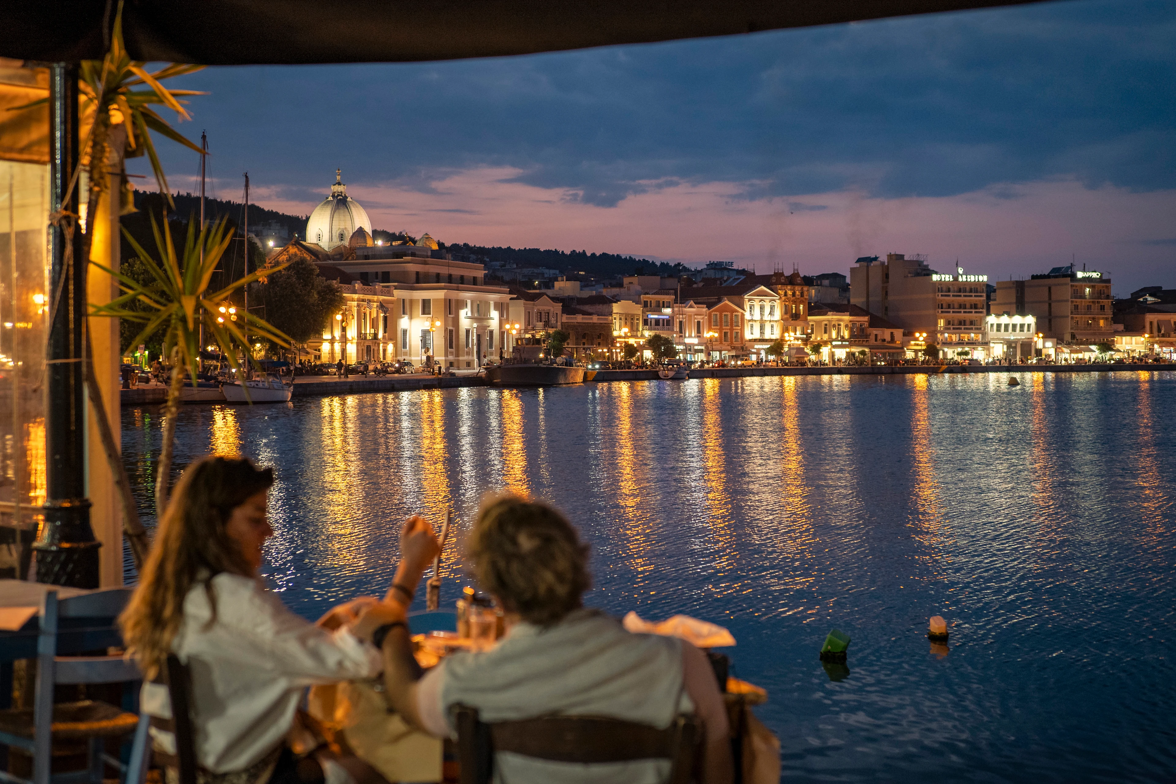 Et par nyter kvelden og utsikten over havnen i Mytilini med opplyste bygninger og restauranter ved vannkanten