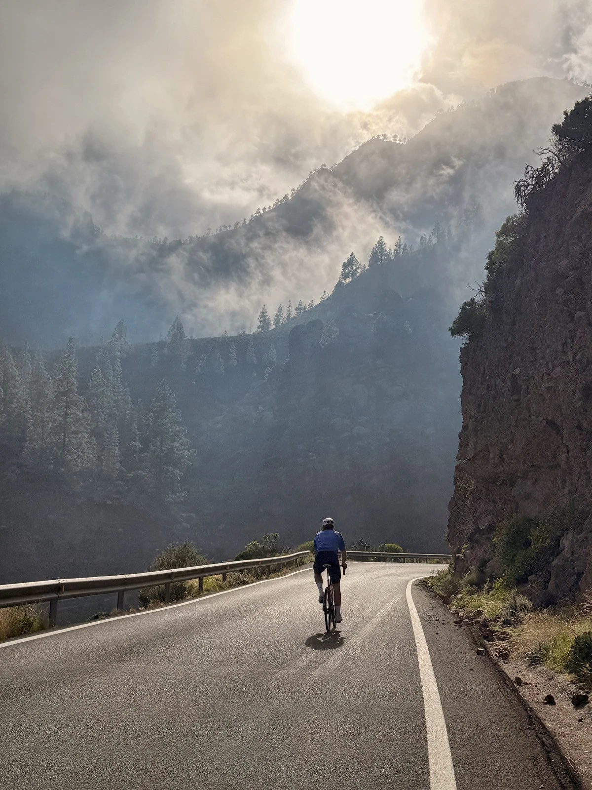 En cyklist färdas uppför en slingrande bergsväg omgiven av höga klippor och dimhöljda tallskogar med solen som bryter igenom molnen ovanför