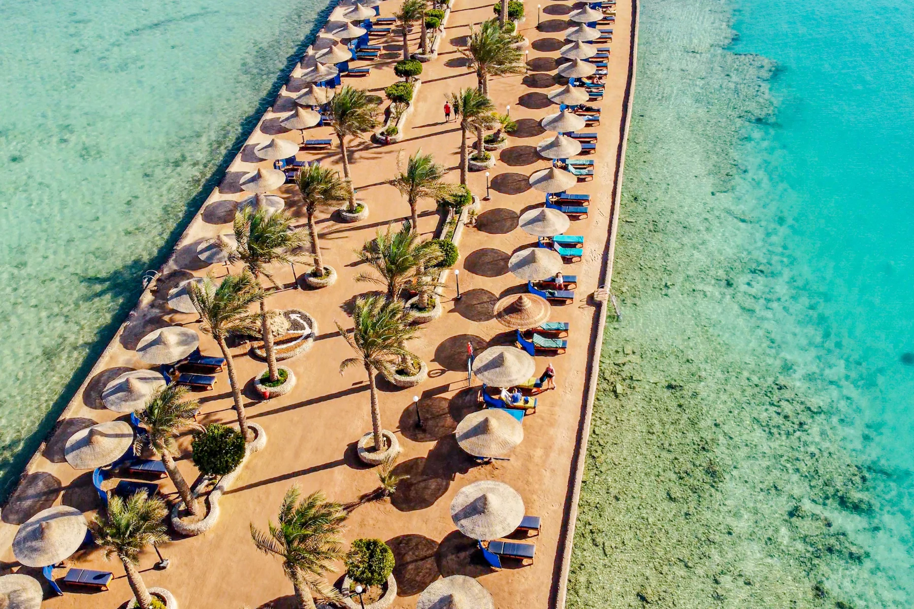 Strandpromenad med solstolar och palmö vid turkost hav, Arabia Azur i Hurghada.
