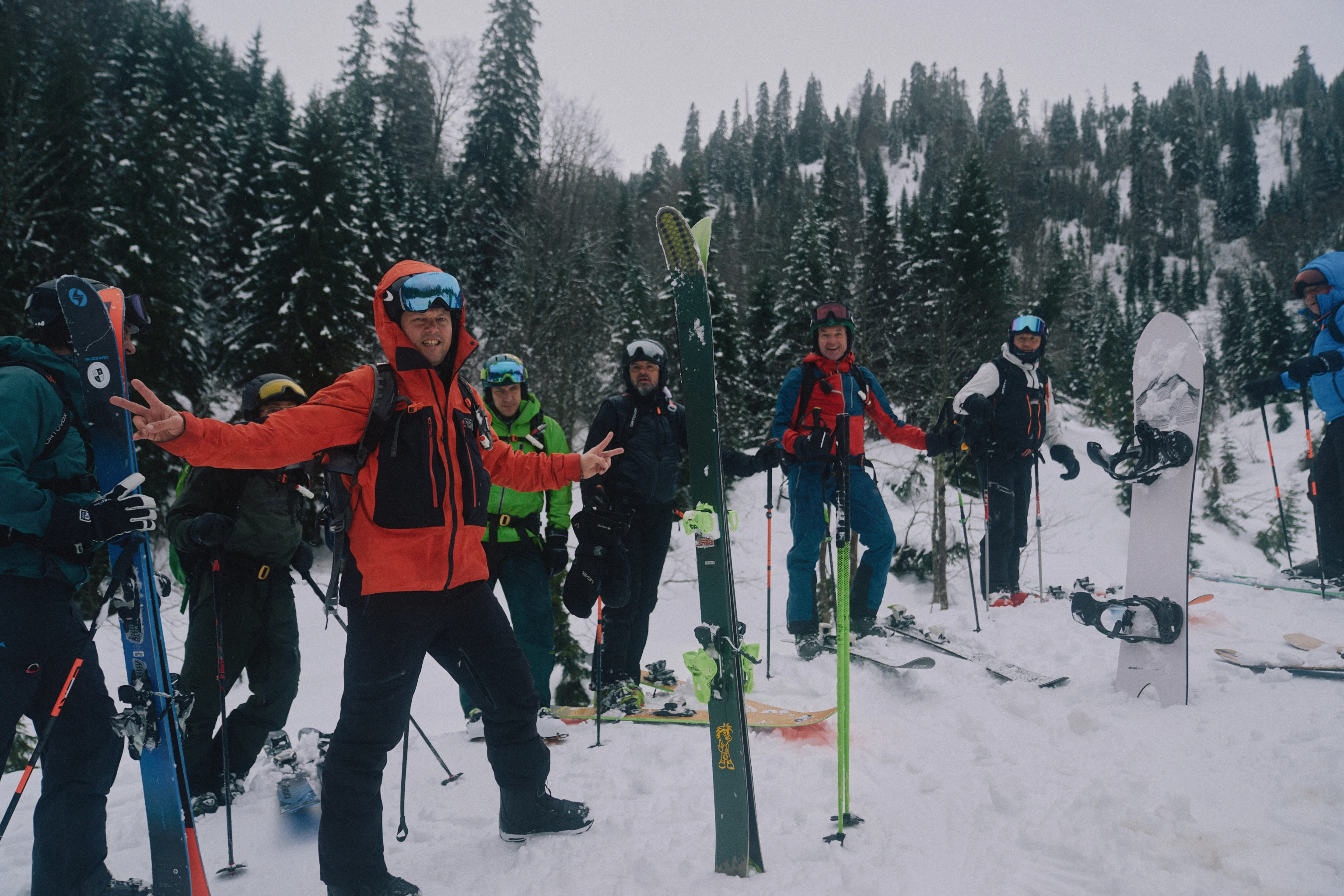Grupp med skid- och snowboardåkare tar paus under en topptur i snötäckt fjällterräng, omgivna av täta granar och vinterlandskap.