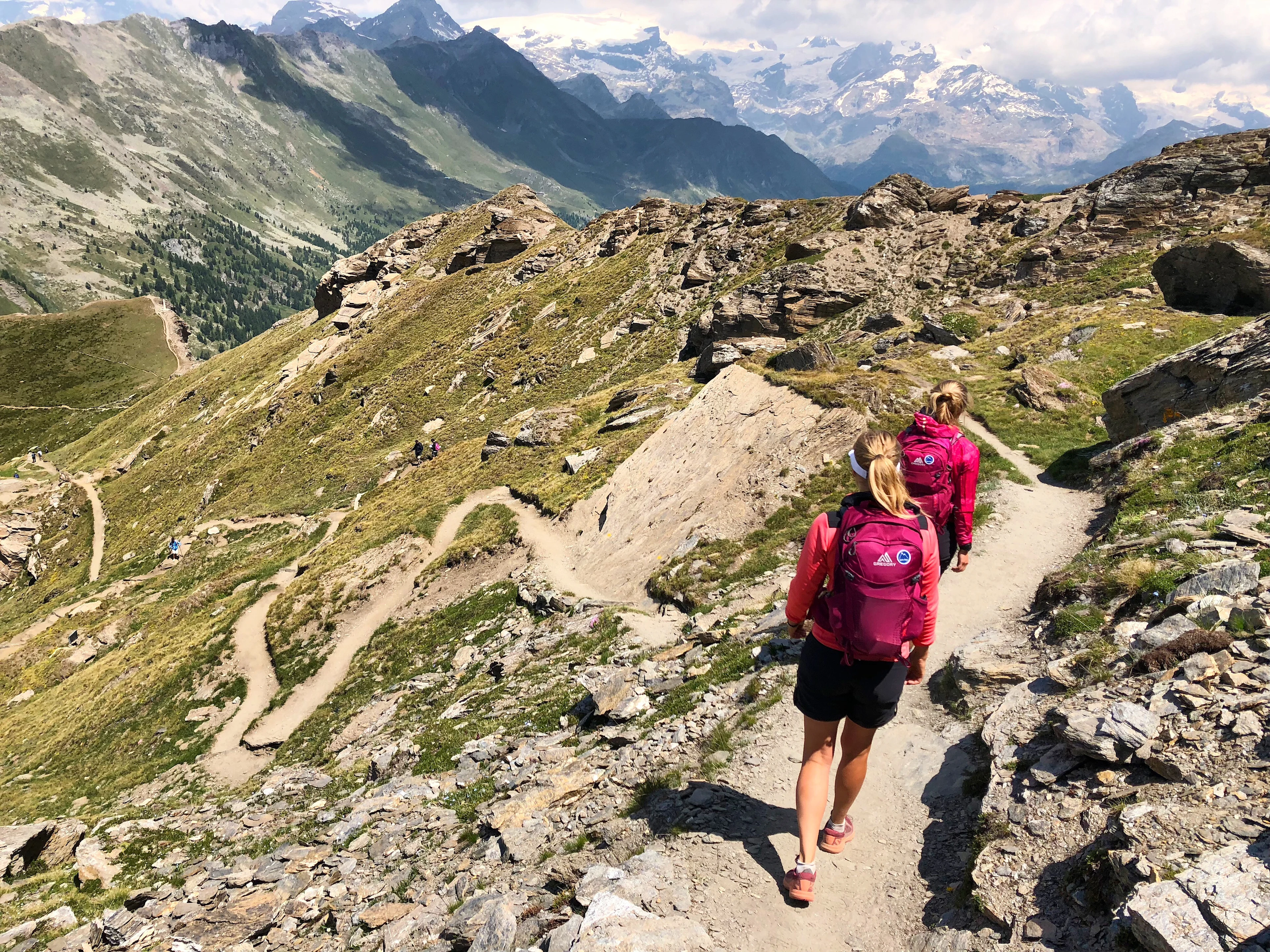 Två kvinnor vandrar längs en bergsstig med utsikt över alpina dalar och snöklädda toppar