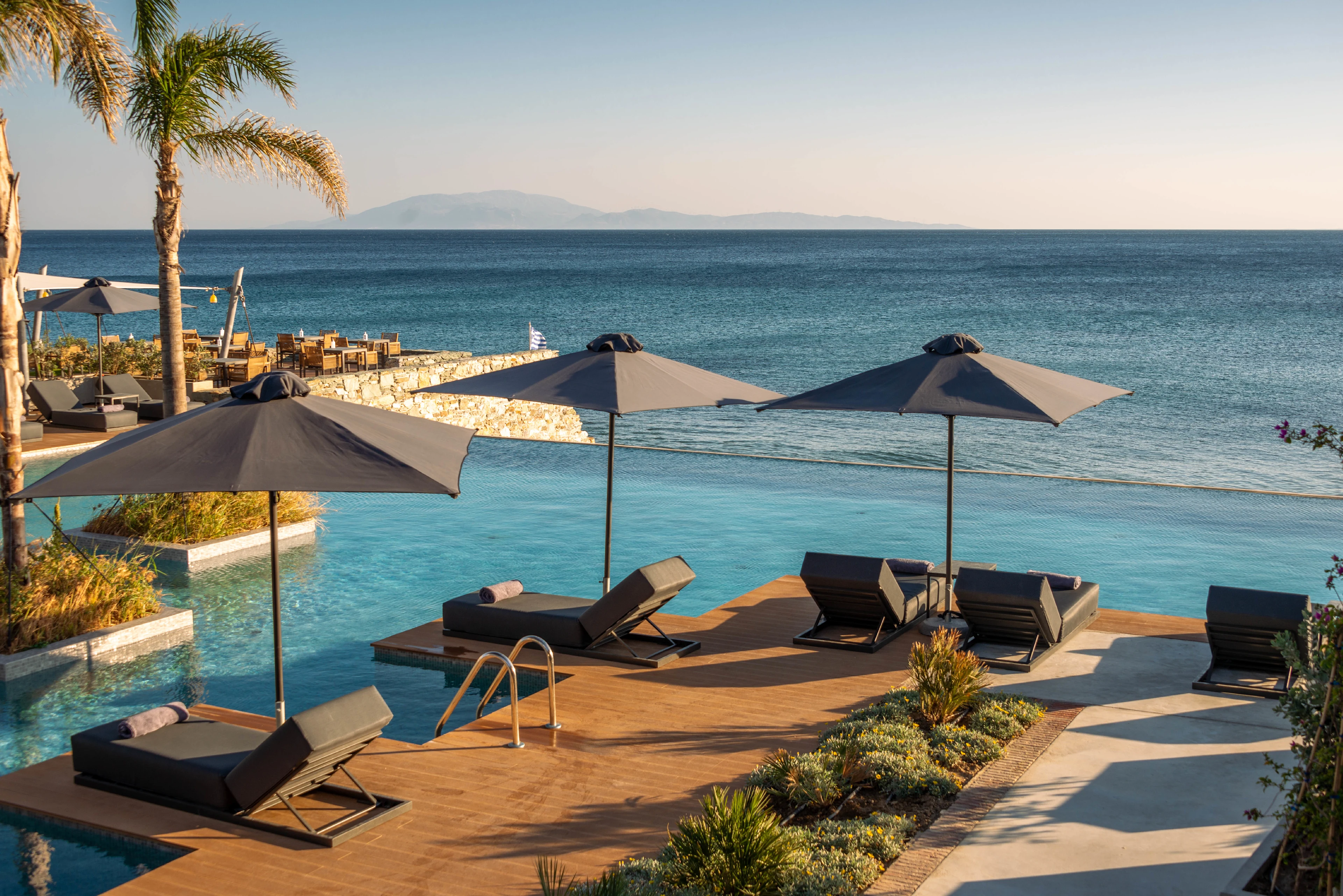 Lyxig infinitypool med solstolar och parasoller vid havet, perfekt för avkoppling med utsikt över bergen i fjärran.