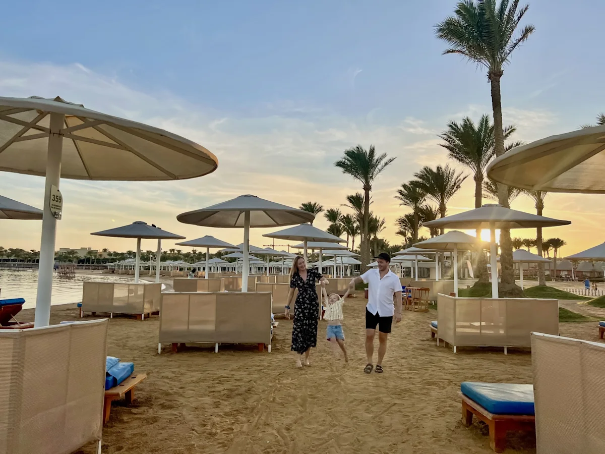 En familj promenerar längs en sandstrand i kvällssol, omgiven av palmer och stora parasoller vid vattnet.