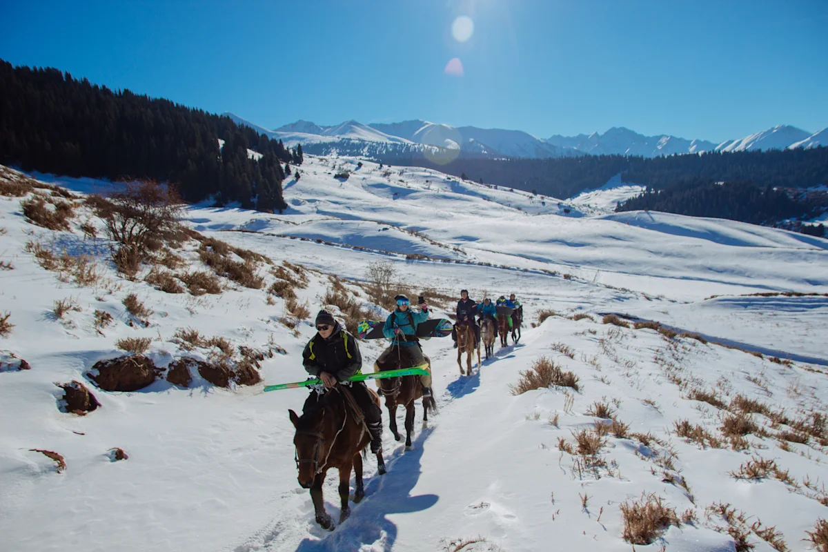 En grupp personer rider på hästar genom ett snötäckt landskap i ett bergsområde under klar himmel.
