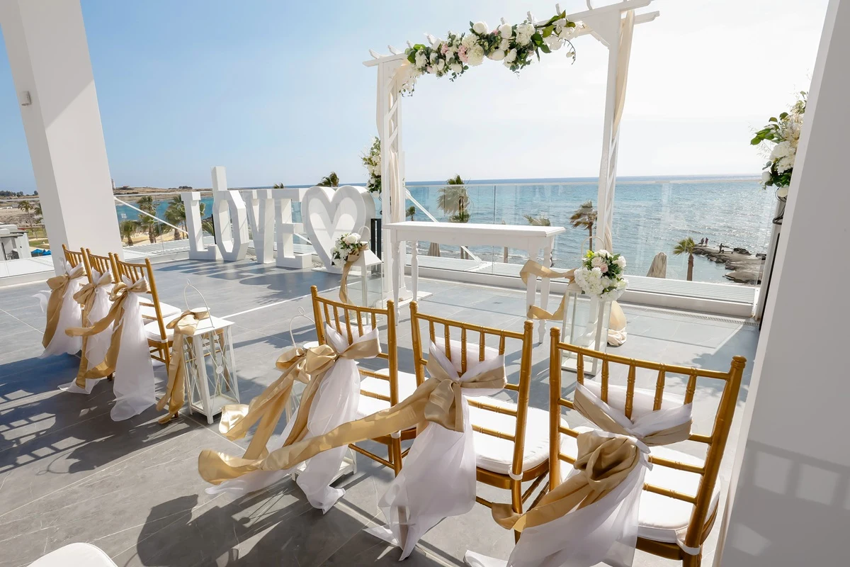 Bryllupoppsetning til en seremoni på terrassen av hotell Bahia Principe Costa Adeje med hvite og gull sløyfer på stolene til gjestene. I bakgrunnen er det skrevet LOVE i hvit, lys skrift med hjerte, bryllupsbue og blått hav og blå himmel i bakgrunnen.