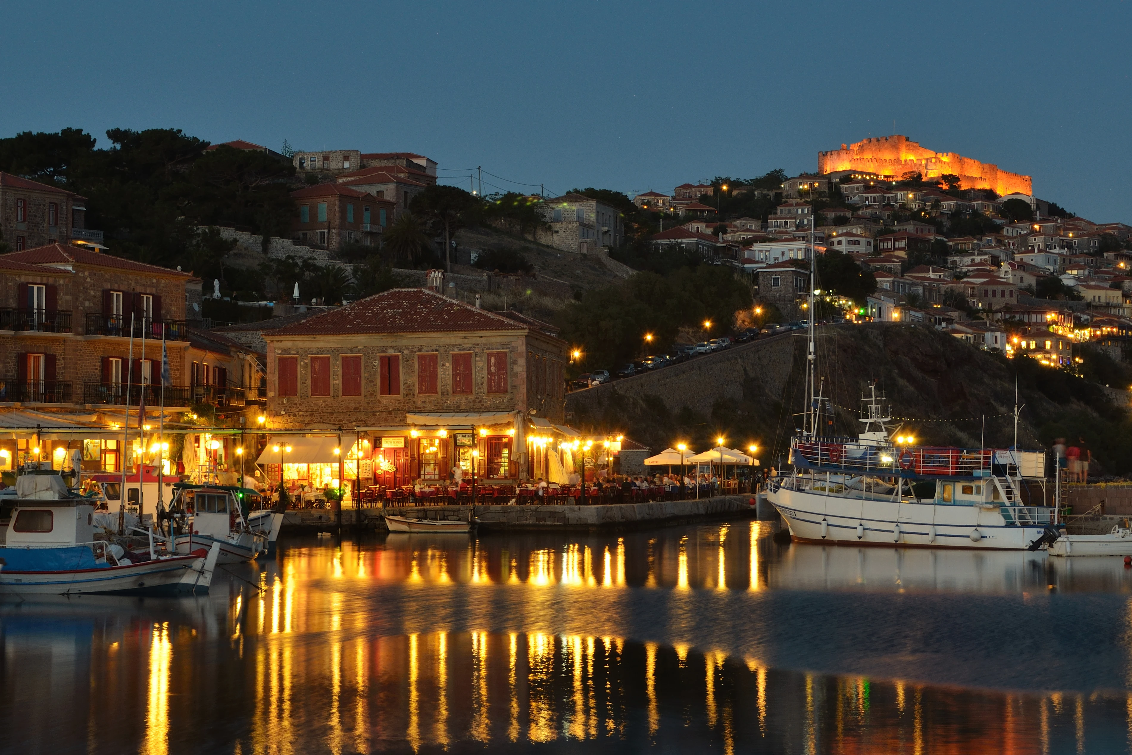 Kvällsbild av Molyvos hamn på Lesvos med upplyst borg, restauranger och båtar i vattnet
