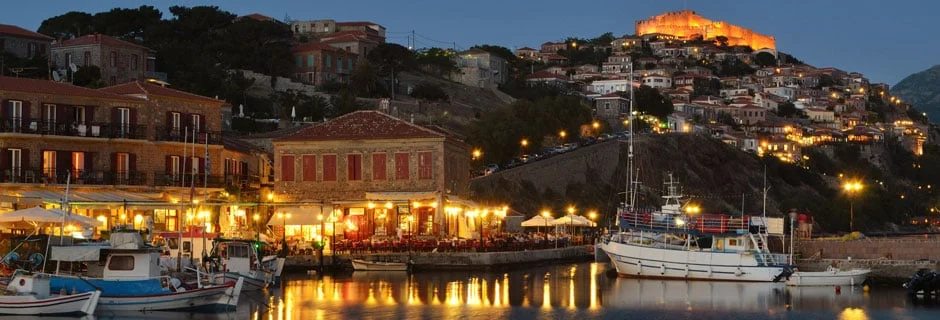 Kveldsutsikt over Molyvos havn på Lesvos med opplyst slott, restauranter og båter i vannet