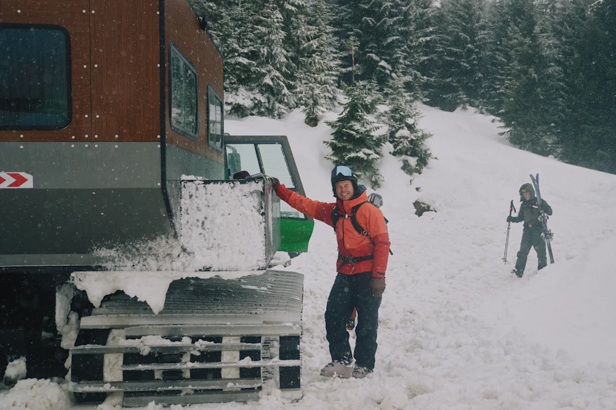 Person i skidutrustning står bredvid bandfordon i snöigt skogslandskap, ytterligare person med skidor i bakgrunden