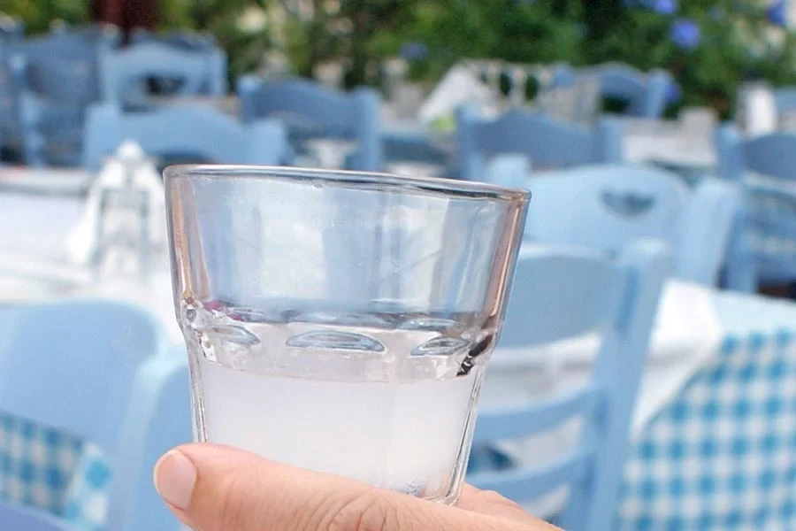 närbild på ett litet glas med ouzo i bakgrunden syns ljusblåa stolar och bord med blåvit-rutiga dukar