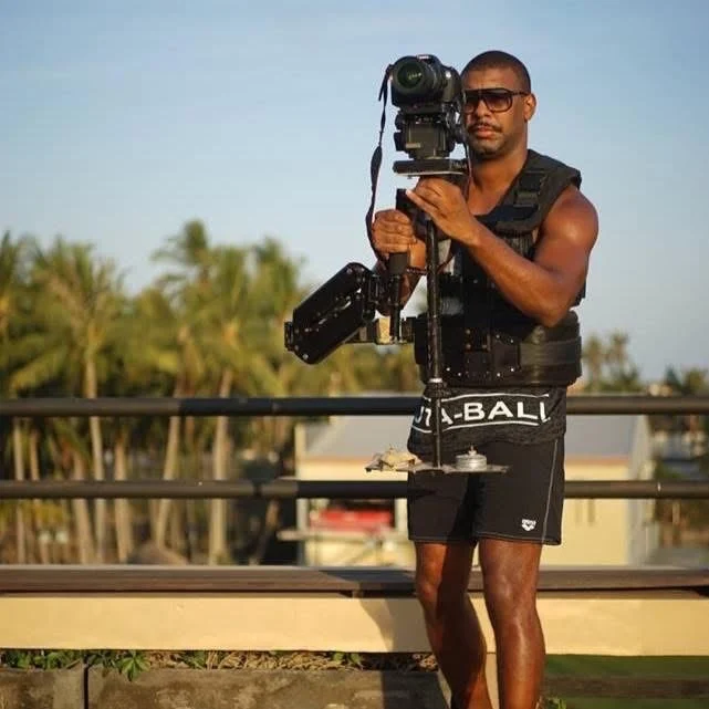 Bilde av Coach David kledd i sort treningssinglet og -shorts som står og filmer ute med palmer i bakgrunnen.